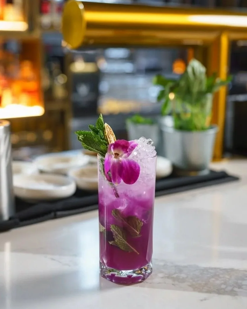 A tall glass with a purple cocktail garnished with pink orchids, mint leaves, and crushed ice on top, set on a marble countertop in a bar or restaurant.