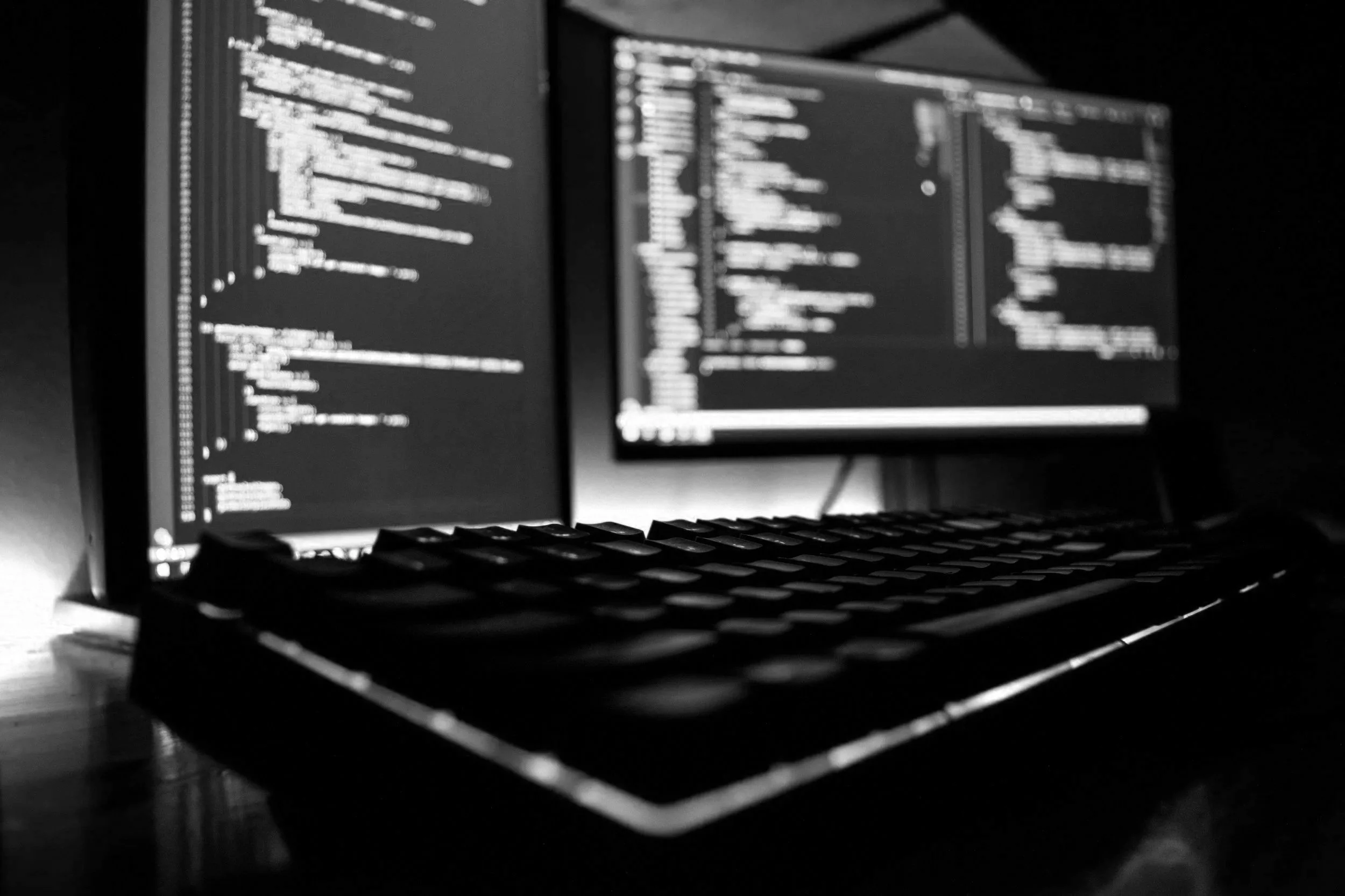A computer workstation with two monitors displaying lines of code, a keyboard in the foreground, and the scene is in black and white.