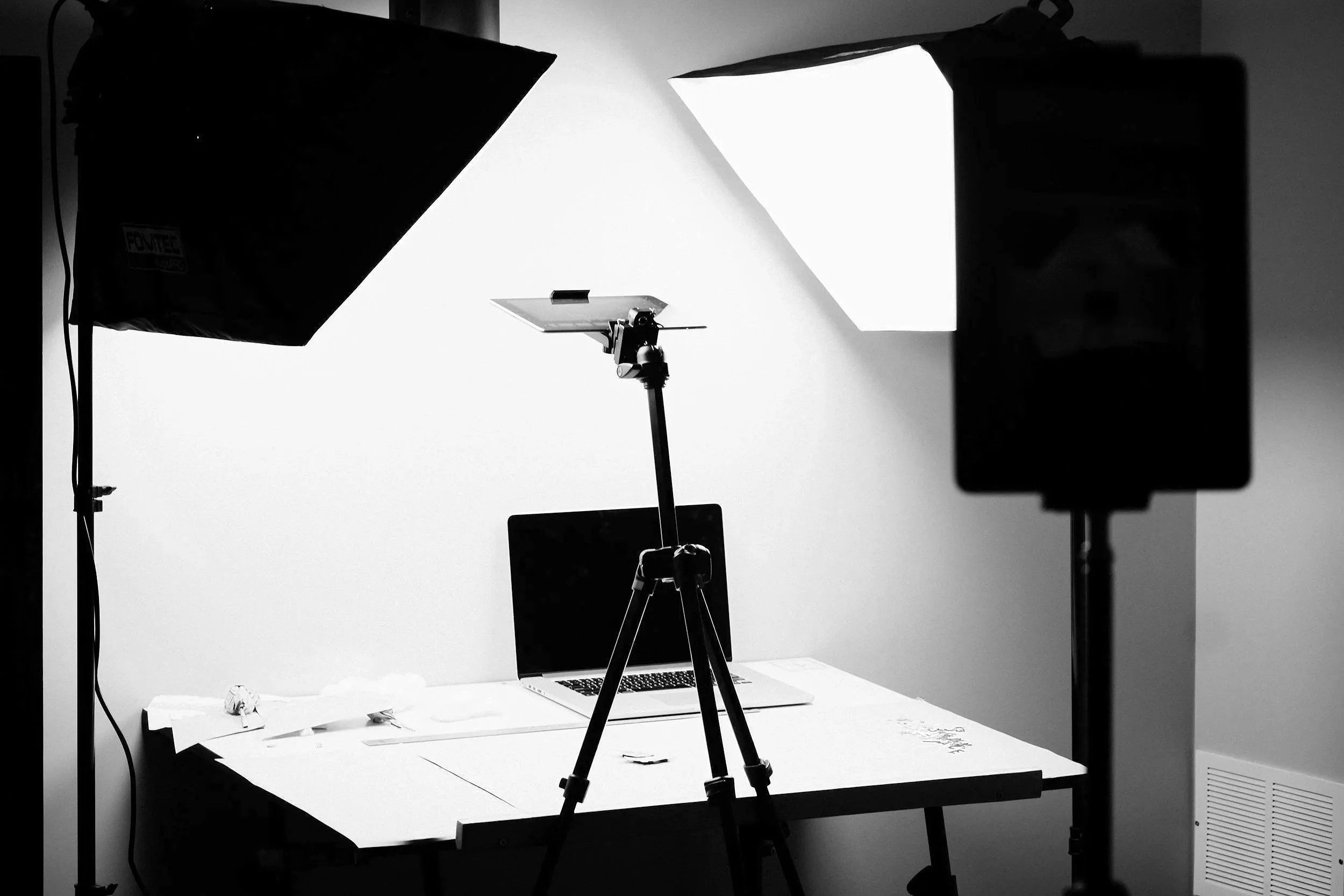 Photography studio setup with two large softbox lights illuminating a white backdrop, a tripod-mounted light, a desk with a laptop, and miscellaneous items on the table.
