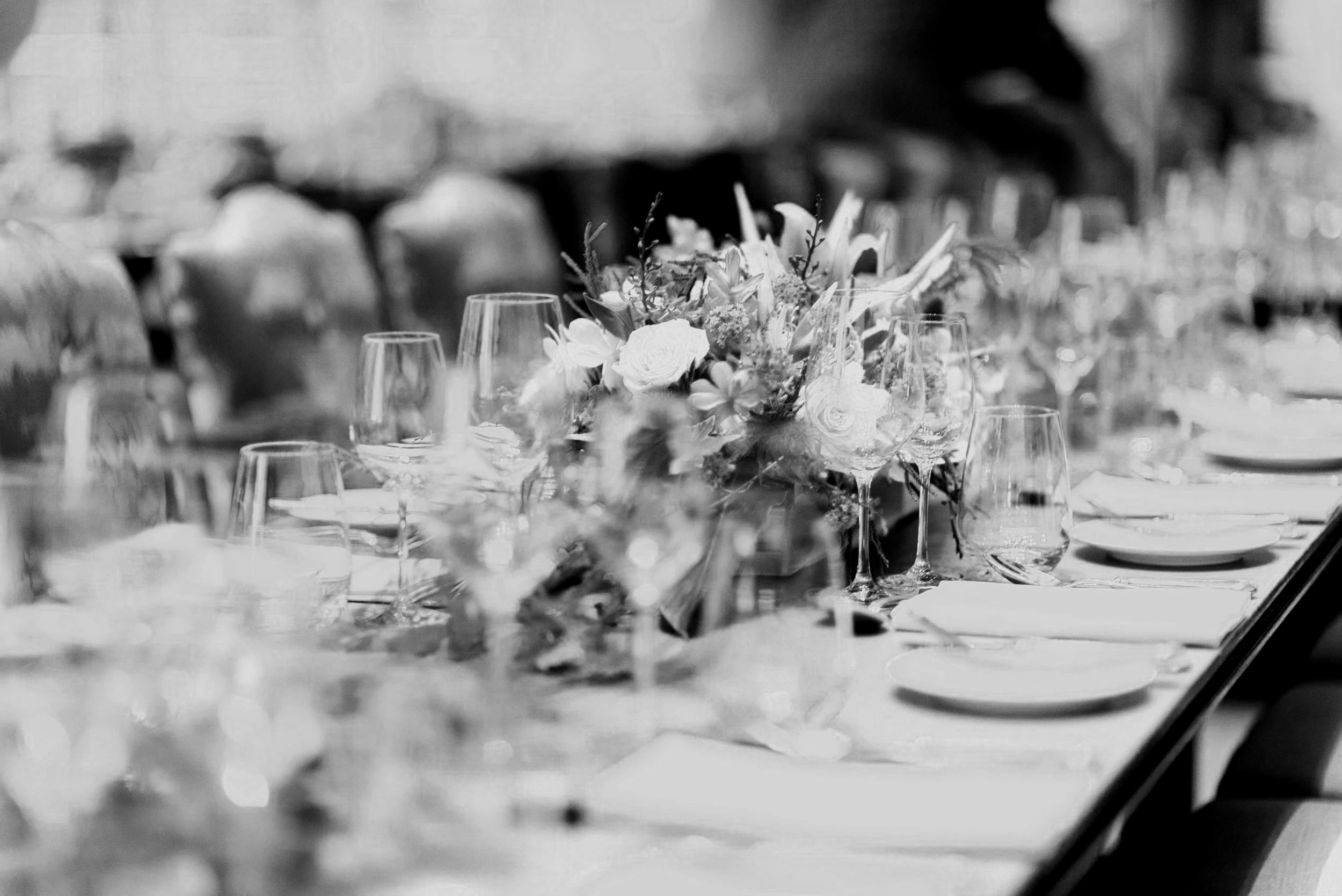 Elegant banquet table set with glassware, plates, and floral centerpieces in a black-and-white photograph.