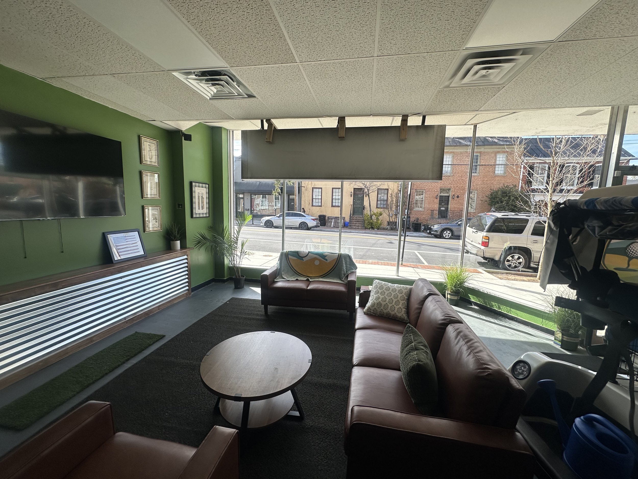 Interior of a cafe or lounge area with green painted walls, a flat-screen TV, framed pictures, a sofa, and a round coffee table. Large windows reveal parked cars and buildings outside, with trees visible in the background.