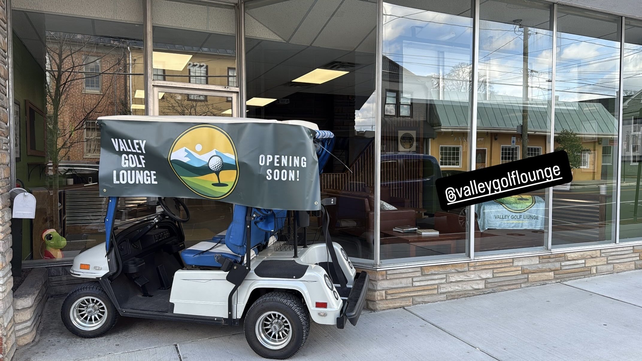 A golf cart parked outside a building, with a sign on the building advertising 'Valley Golf Lounge Opening Soon!' and a towel with the same logo inside. The building has large glass windows, and reflections of nearby houses and power lines are visible.