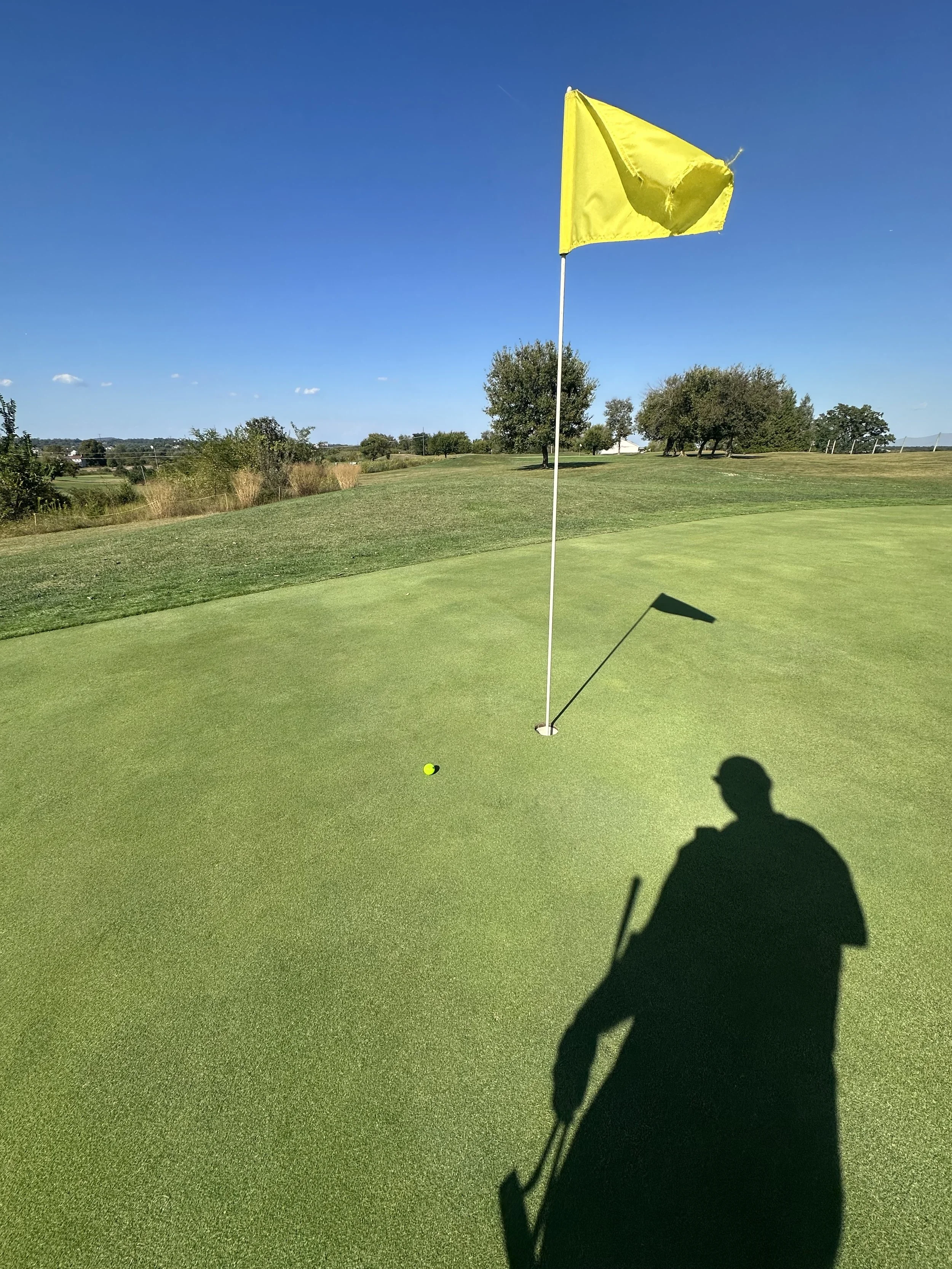 A yellow golf flag on a green golf course, with a golf ball nearby and a person's shadow on the ground, under a clear blue sky.