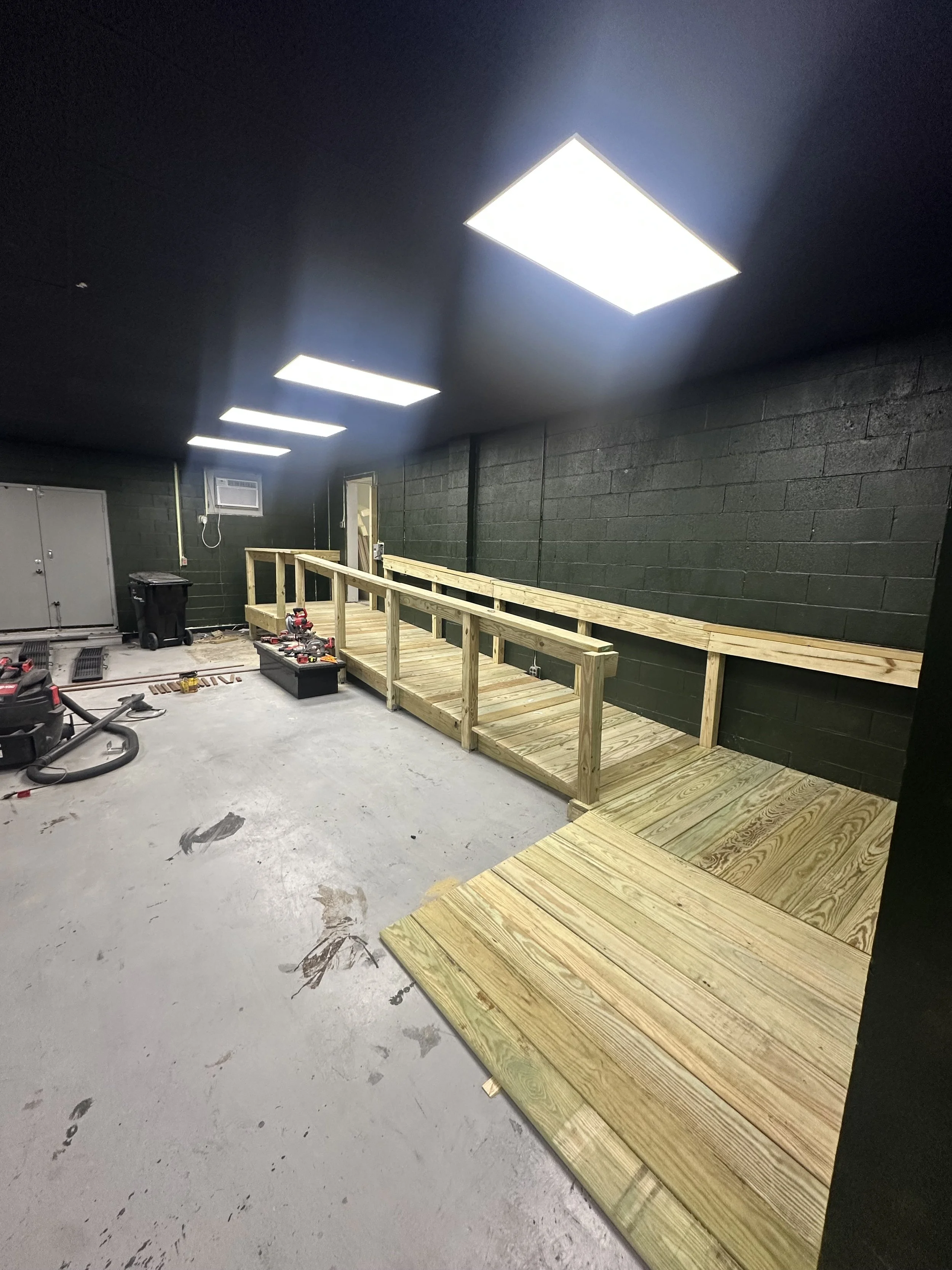 Construction site with black painted cinder block wall, ceiling lights, a small wooden ramp, and construction tools and materials on the floor.