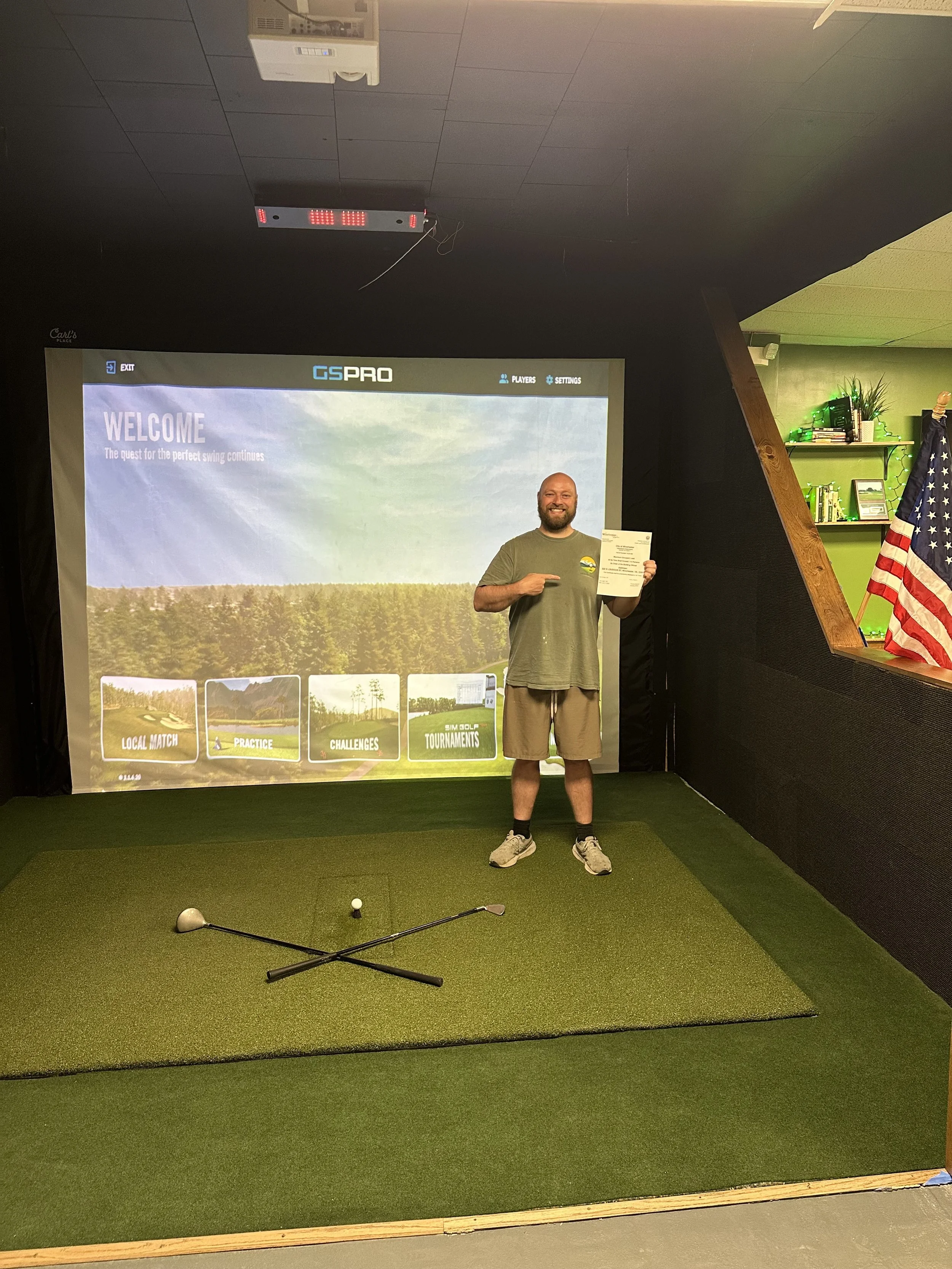 A man standing on a green indoor golf putting mat, holding a certificate and pointing at it, with a golf club lying on the mat. Behind him is a screen with golf game options, an American flag to the right, and a shelf with books and decorations.