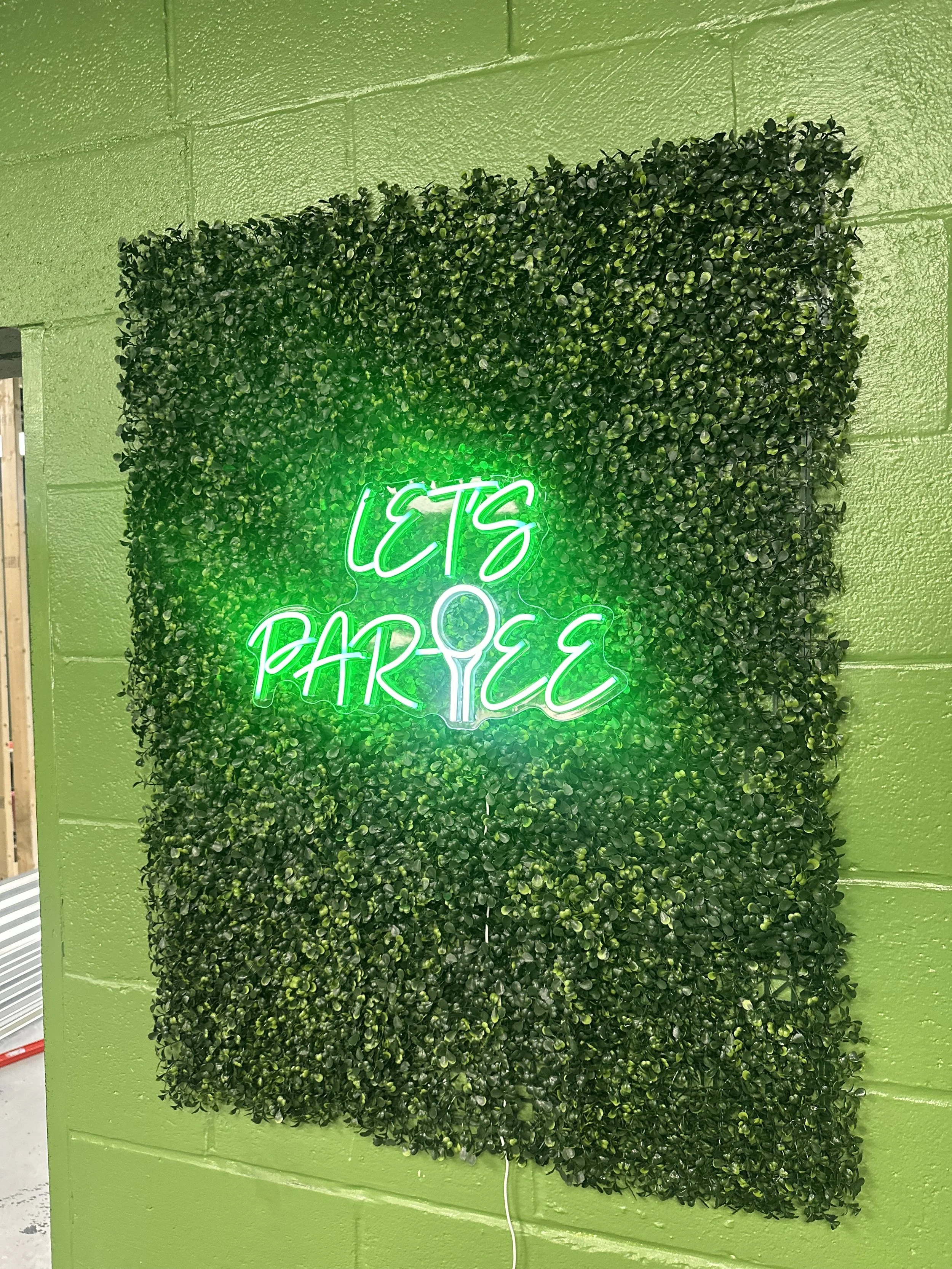 Green leafy wall with neon sign reading 'Let's Par-te'