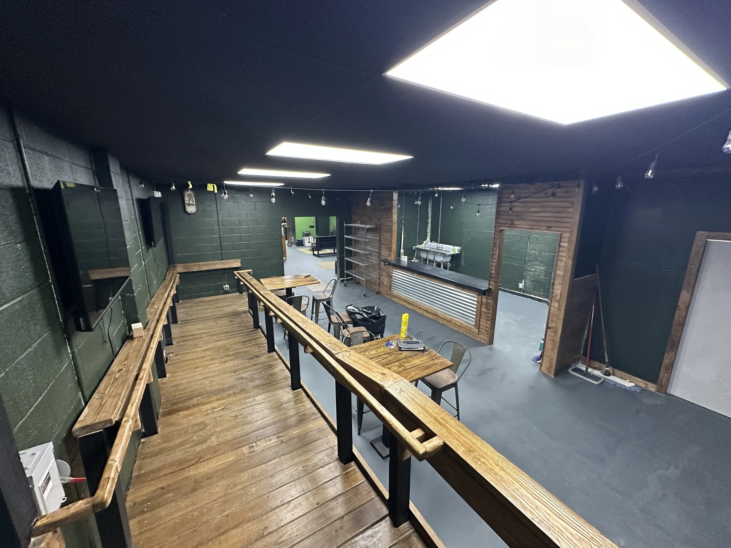 A room with dark walls and ceiling, illuminated by large rectangular ceiling lights. There is a wooden bar counter with matching wooden benches and tables along a railing, facing a smaller area with shelves and a sink. Some chairs and miscellaneous items are scattered around.