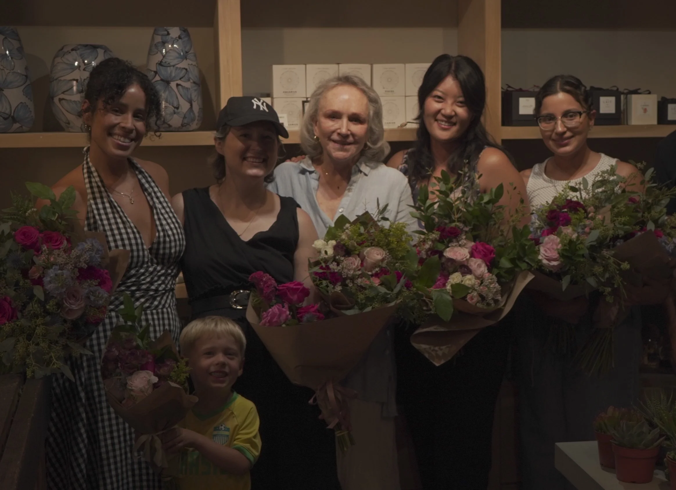 Group of women and a young boy holding large bouquets of flowers, celebrating indoors with shelves of boxes and decorative vases in the background.