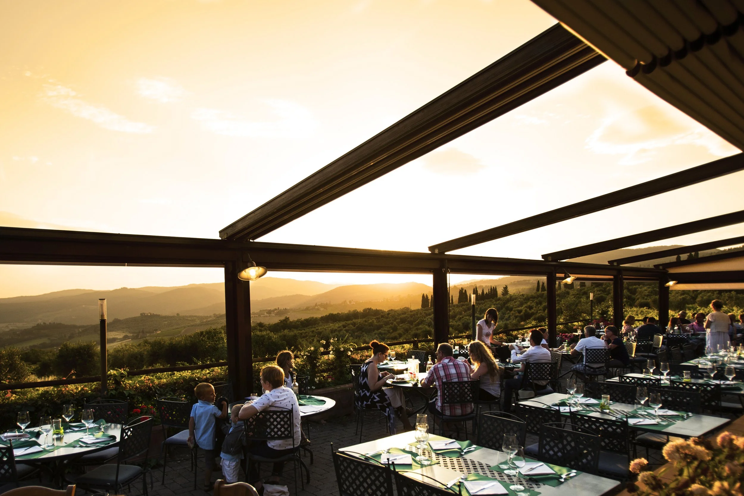 Outdoor restaurant patio with people dining at tables during sunset, hillside landscape in the background.