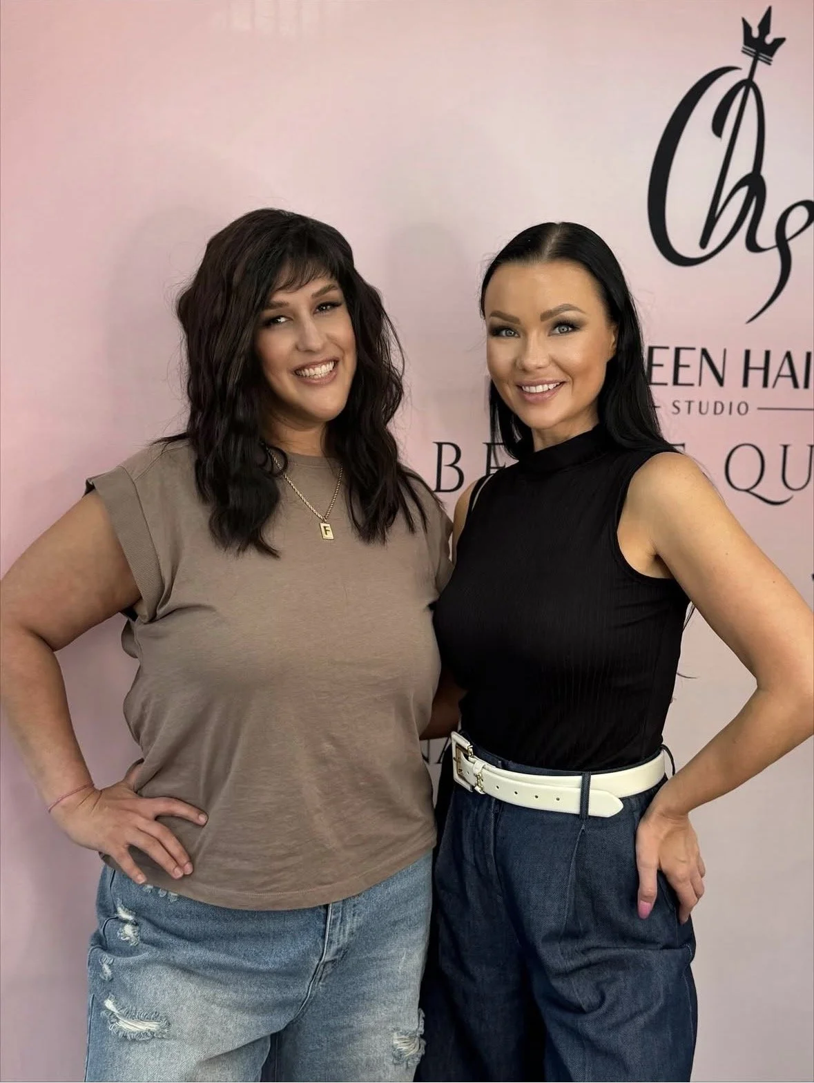 Two women posing together in front of a pink backdrop with black text logo for a hair salon. One woman is wearing a tan t-shirt and ripped jeans, the other is in a black sleeveless top and dark jeans with a white belt.
