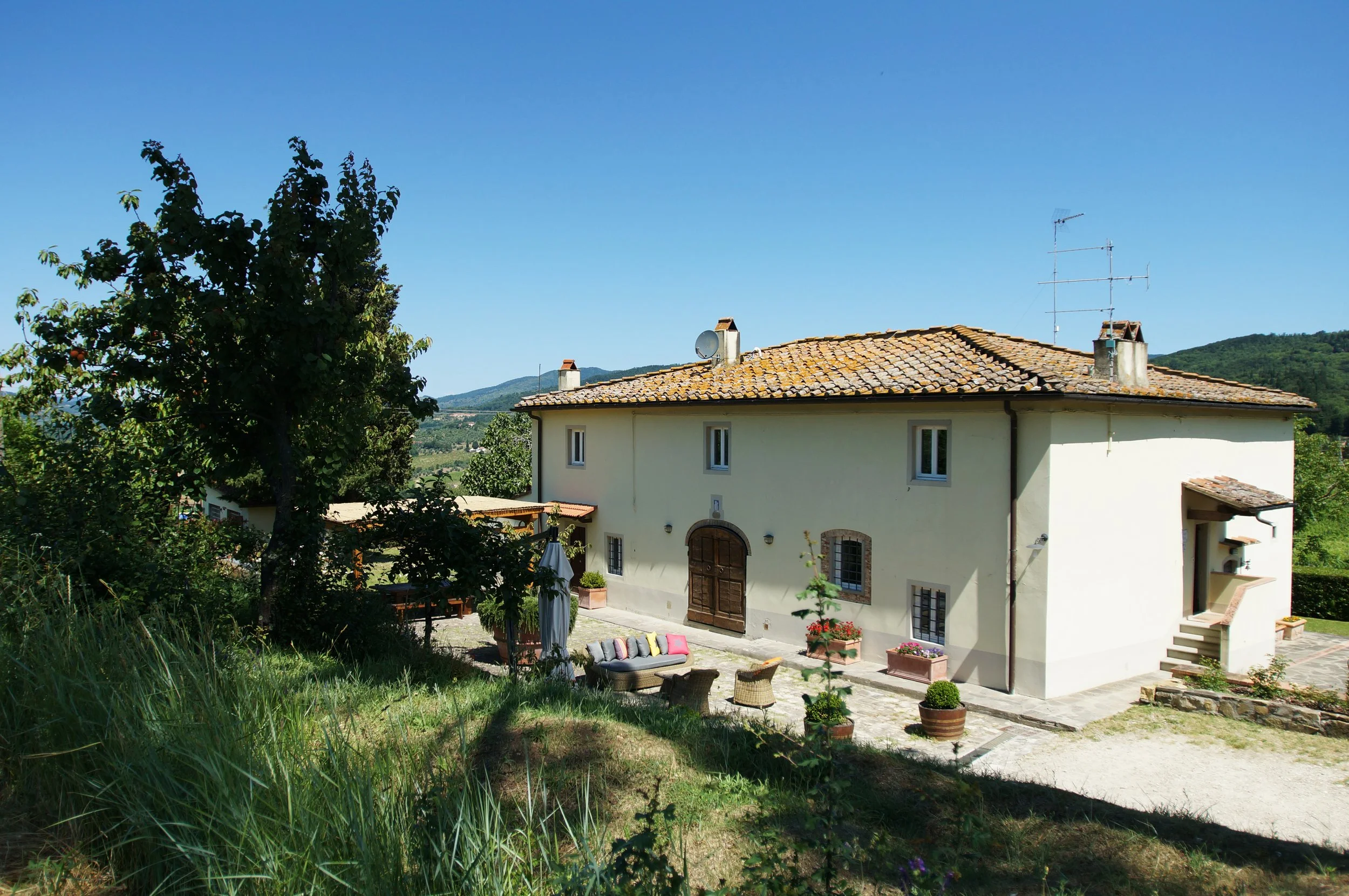 A two-story house with a tiled roof, surrounded by trees and a garden with outdoor furniture, potted plants, and a clear blue sky in a rural area.