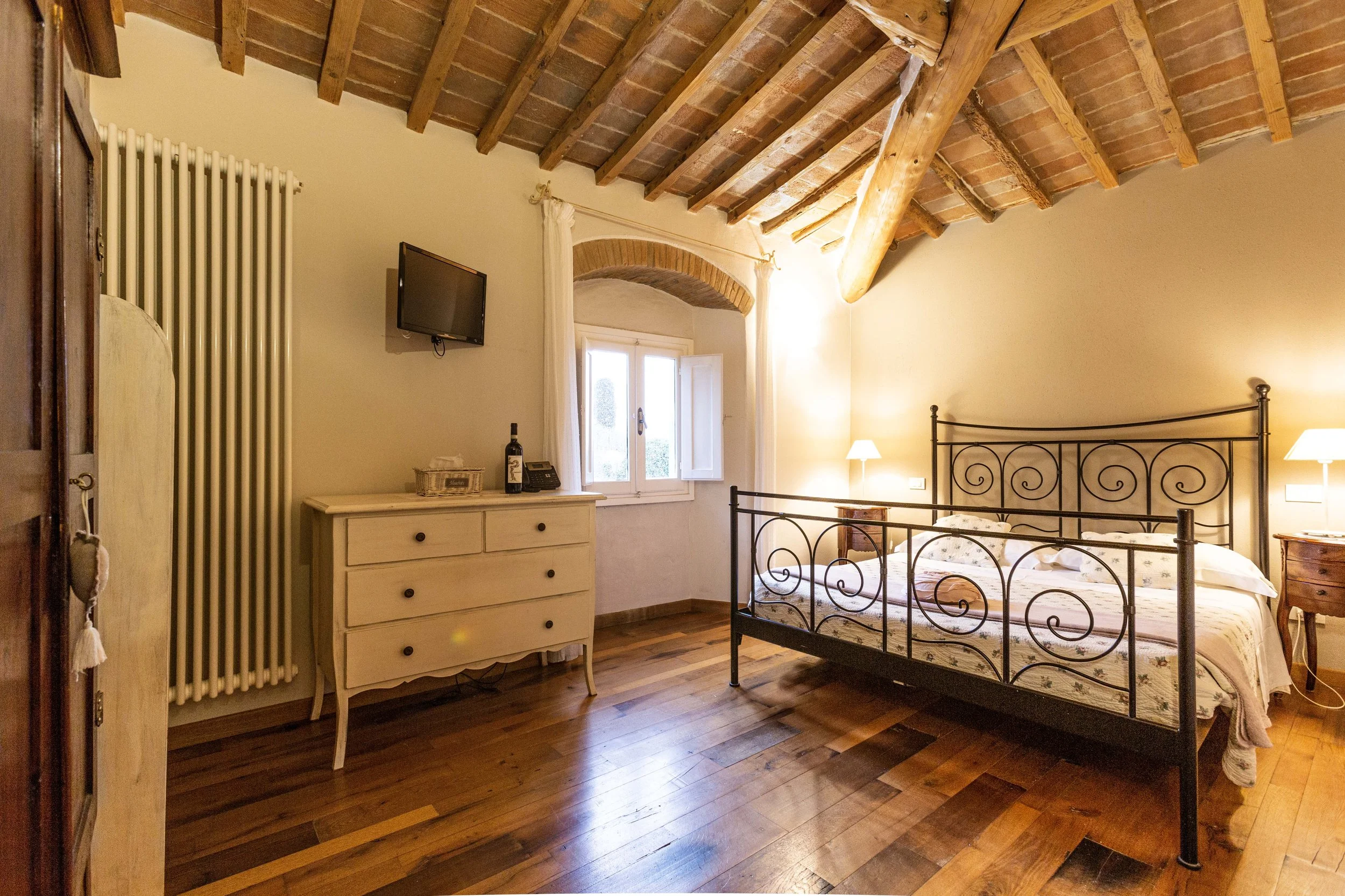 A cozy bedroom with a wooden ceiling, metal bed frame, small nightstands with lamps, a window with curtains, a dresser, and a small TV on the wall.