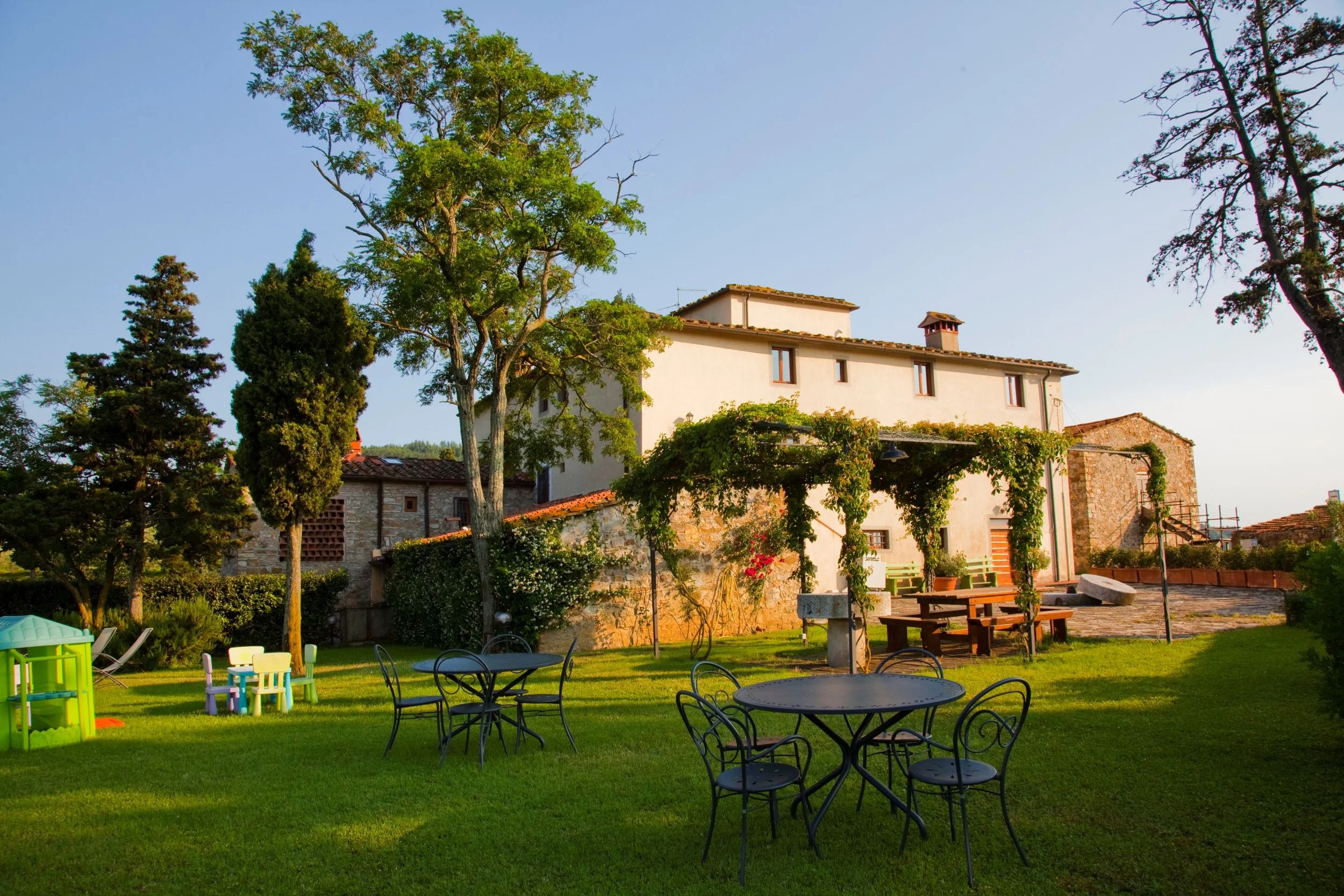 A backyard garden with green grass, outdoor tables and chairs, surrounded by large trees, with a house and clear sky in the background.