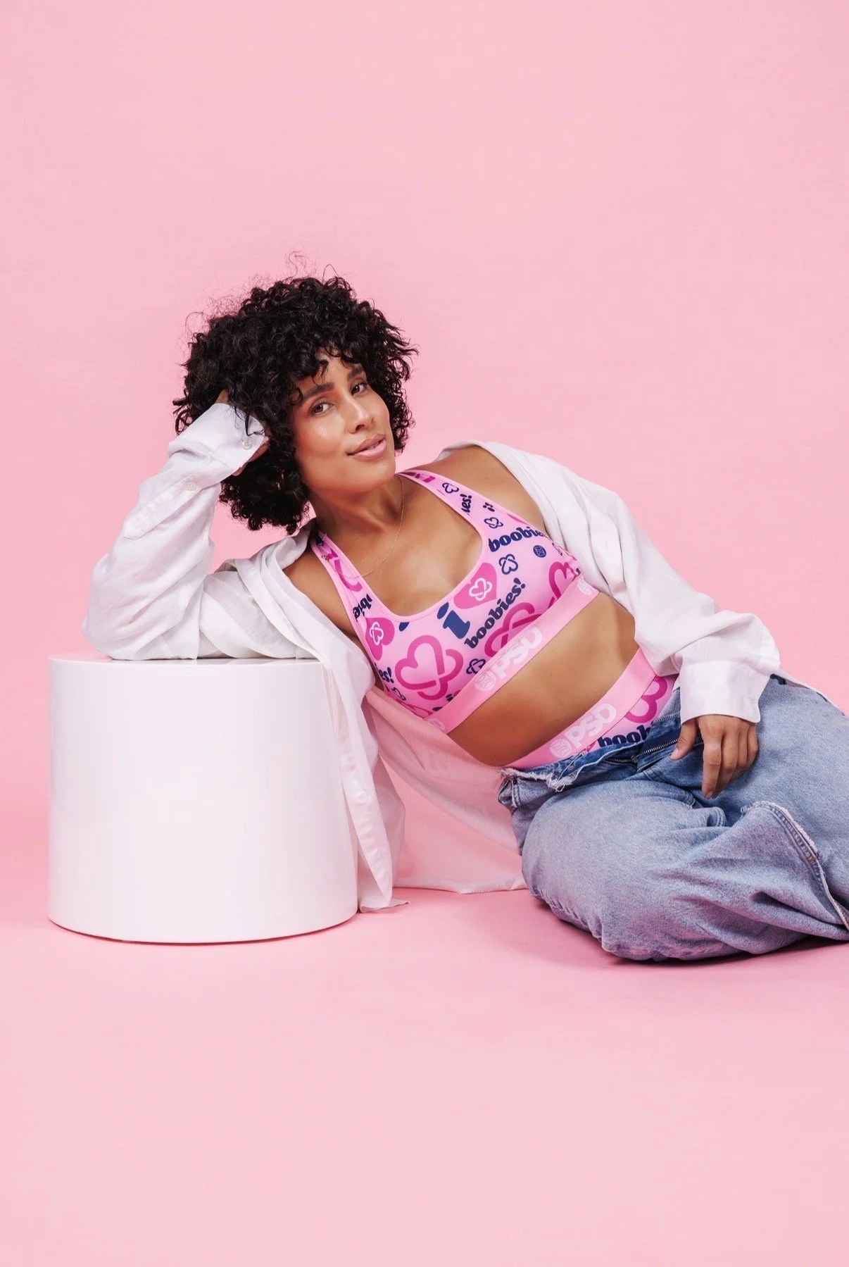 A woman with curly dark hair lying on a pink background, resting her head on her hand on a white cylindrical table, wearing a colorful printed sports bra and loose jeans, with a white unbuttoned shirt.