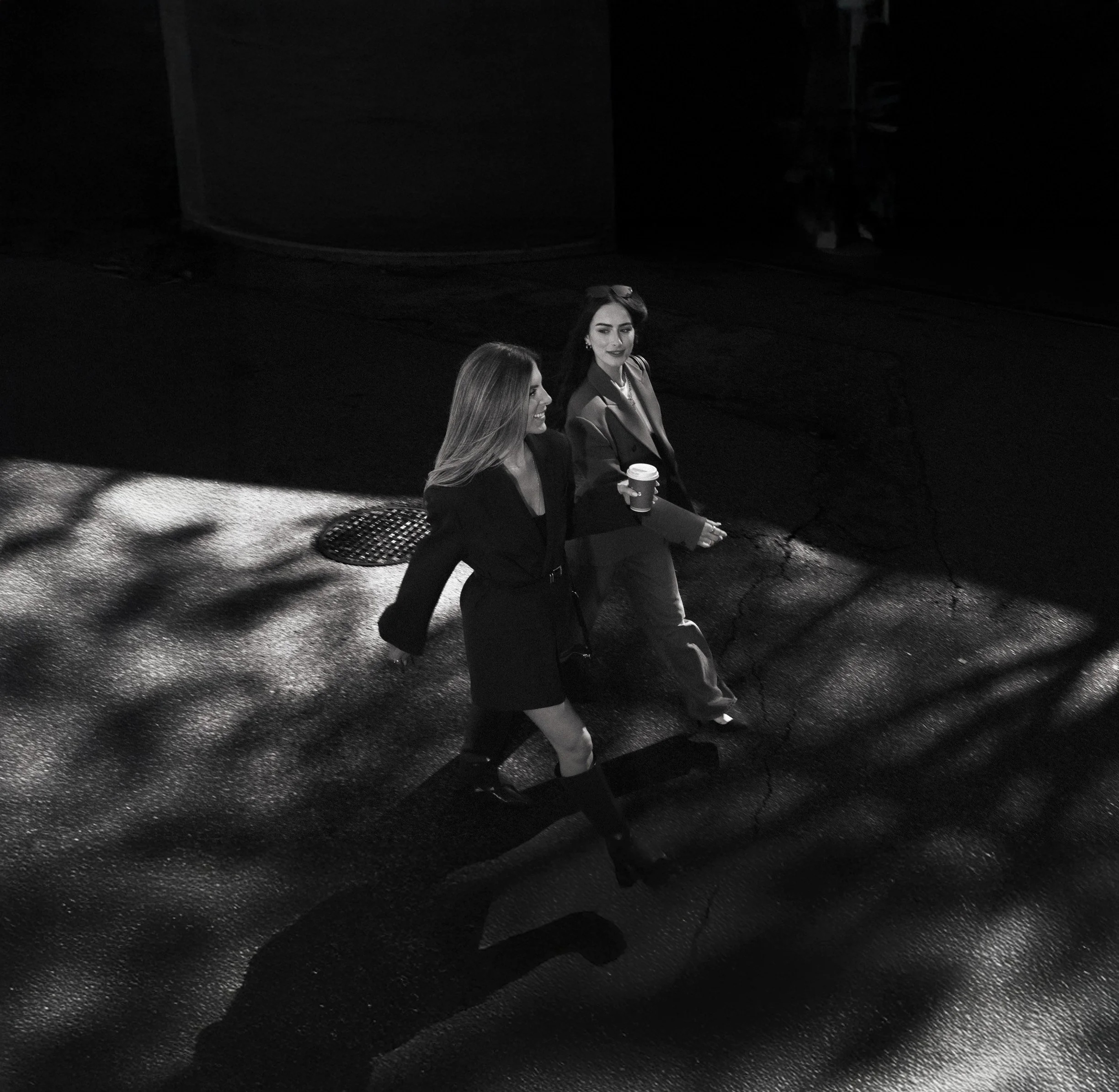 Two women walking on city street at night, one holding a coffee cup, with shadows of trees falling on the pavement, viewed from above.