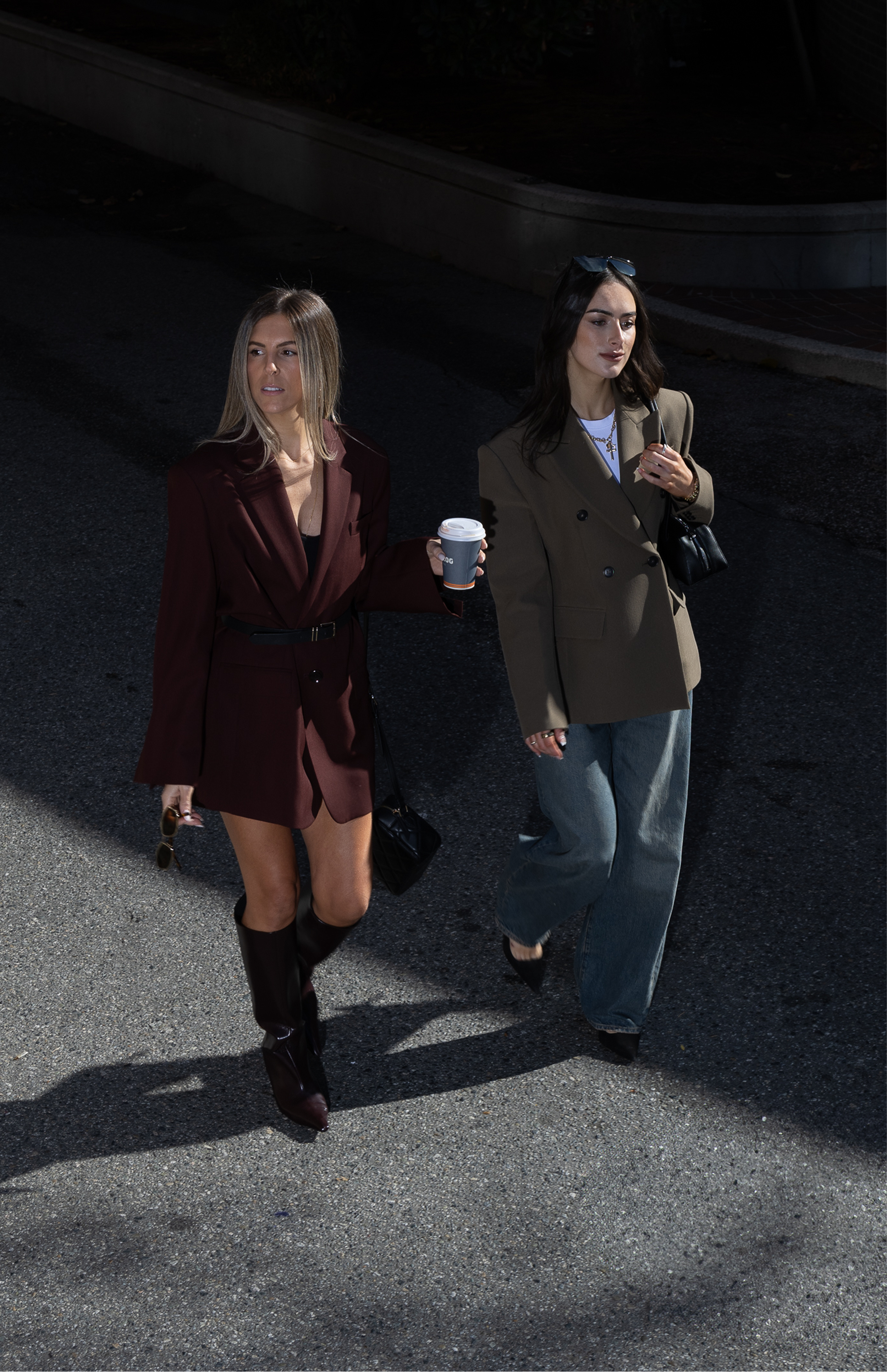 Two women walking on a city street at night, one wearing a blazer and jeans, the other in a blazer dress and boots, holding coffee cups and sunglasses.