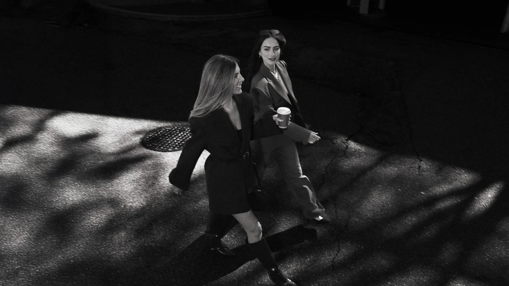 Two women walking on a dark street at night, one holding a coffee cup, shadows cast by nearby trees