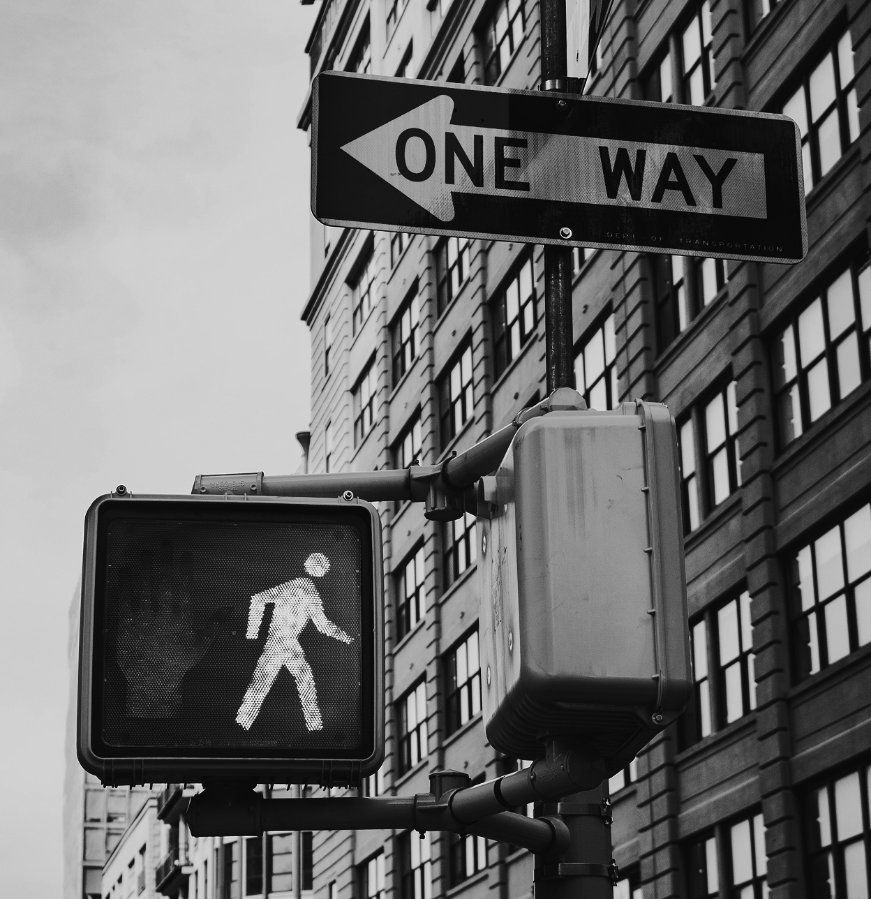 Black and white photo of a pedestrian traffic light with a green walking figure, a one-way street sign pointing left, and a city building in the background.