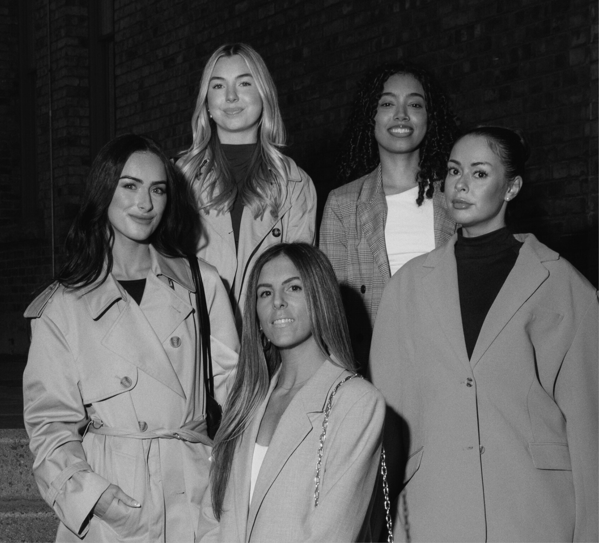 Six women standing outdoors against a brick wall, wearing fashionable trench coats and blazers, looking at the camera.