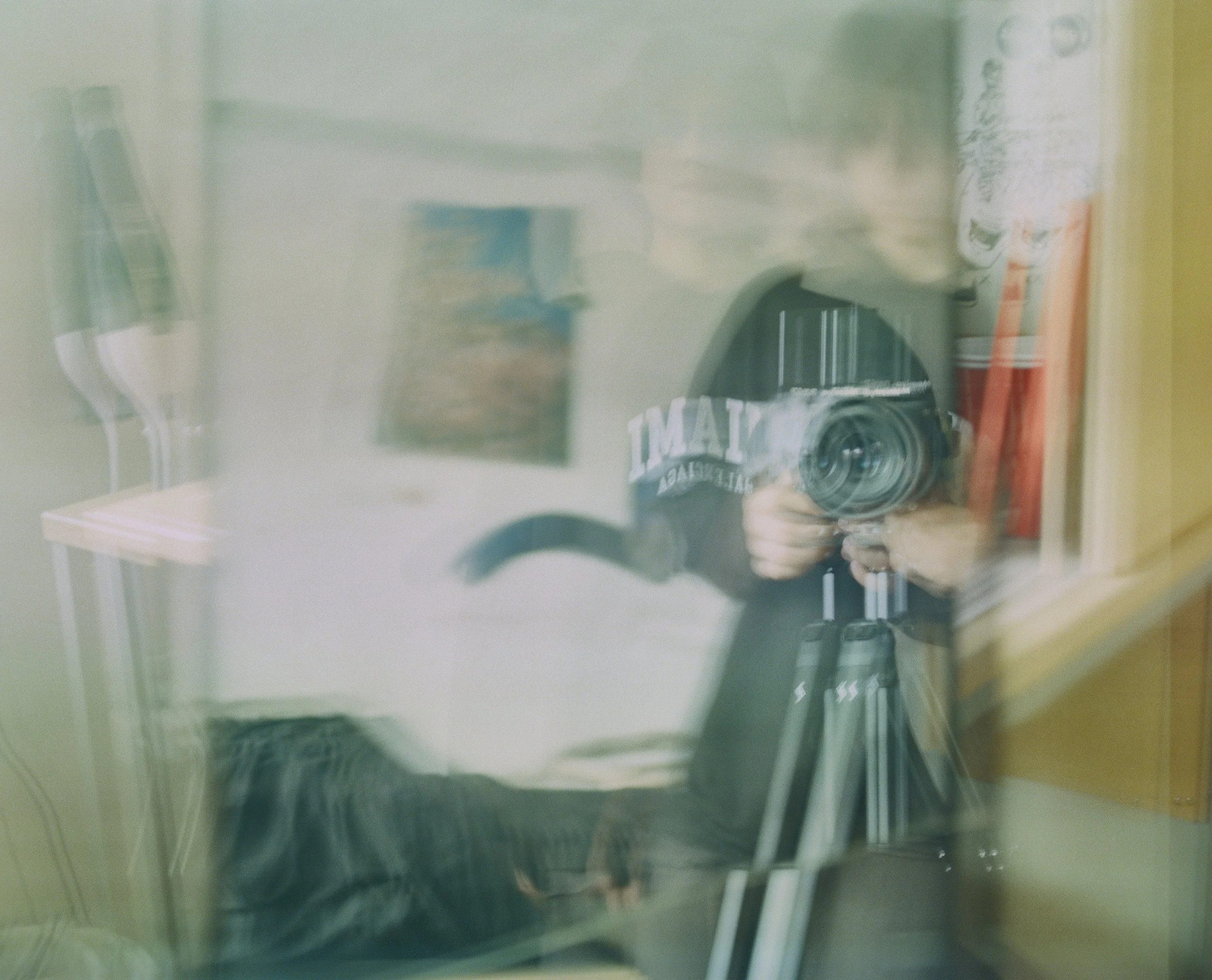 Reflection of a person taking a photograph with a camera in a glass surface, with various objects including books and furniture visible behind.