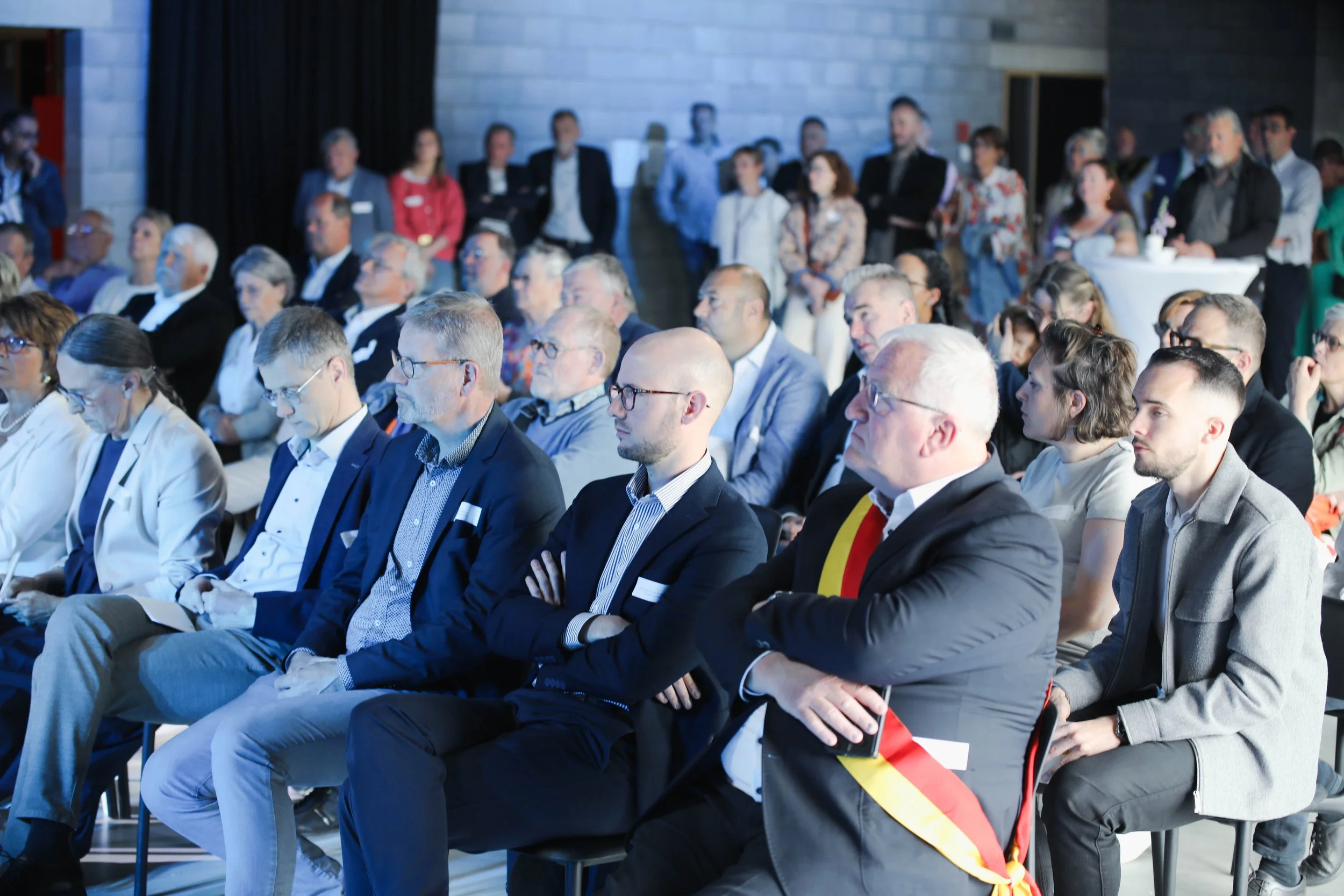 Une audience assise lors d'une conférence ou d'un événement officiel au hall de Clavier, avec plusieurs personnes portant des tenues professionnelles et certains en costumes, dont un homme avec une écharpe ou une chaîne tricolore, dans un espace inté