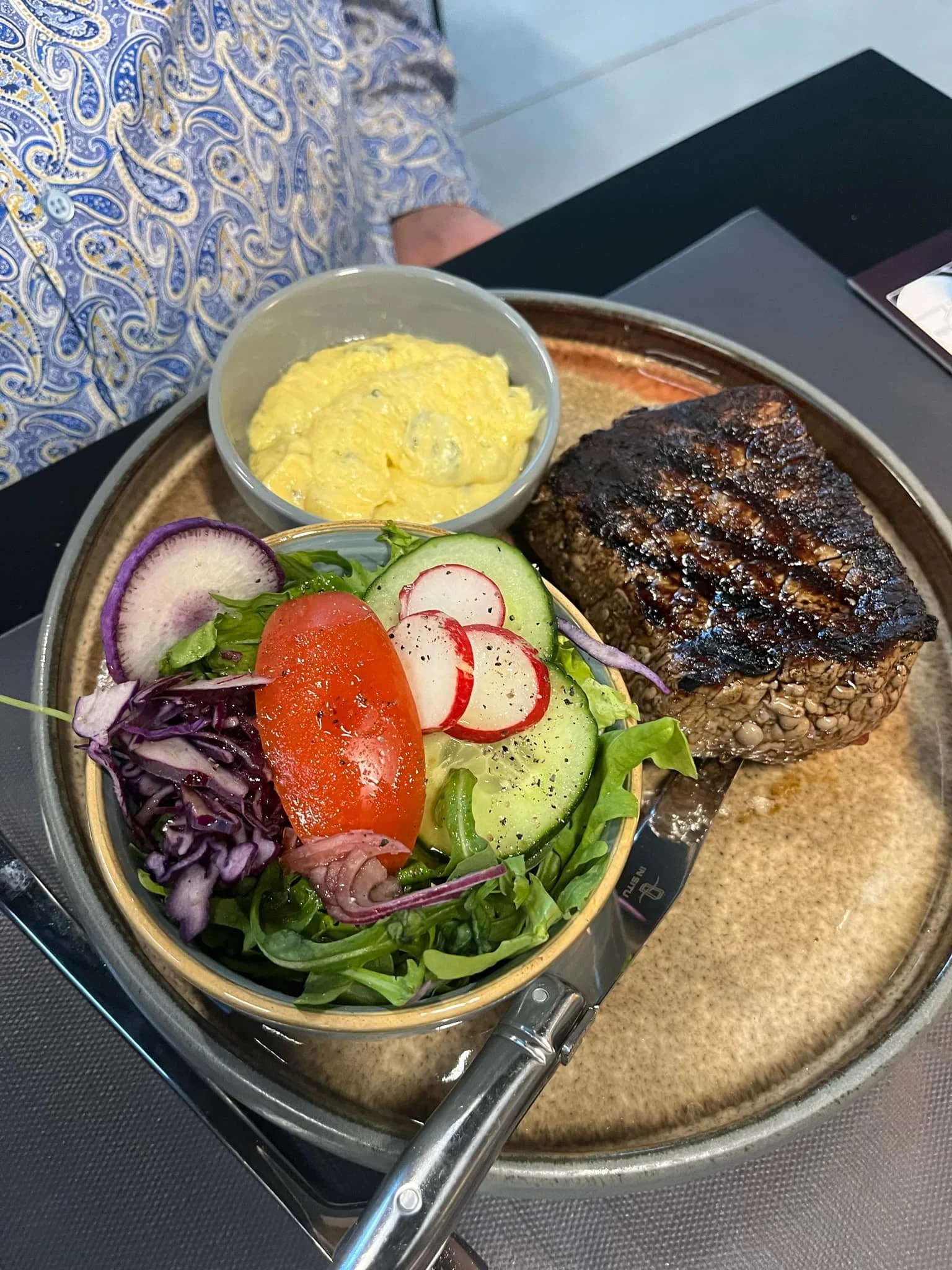 Un plat comprenant un steak grillé, une salade variée avec des tomates, concombres, radis, et chou rouge, un bol de purée d'œufs, et une sauce ou sauce. Il est servi sur une assiette en céramique sur une table  - Aftersport Clavier