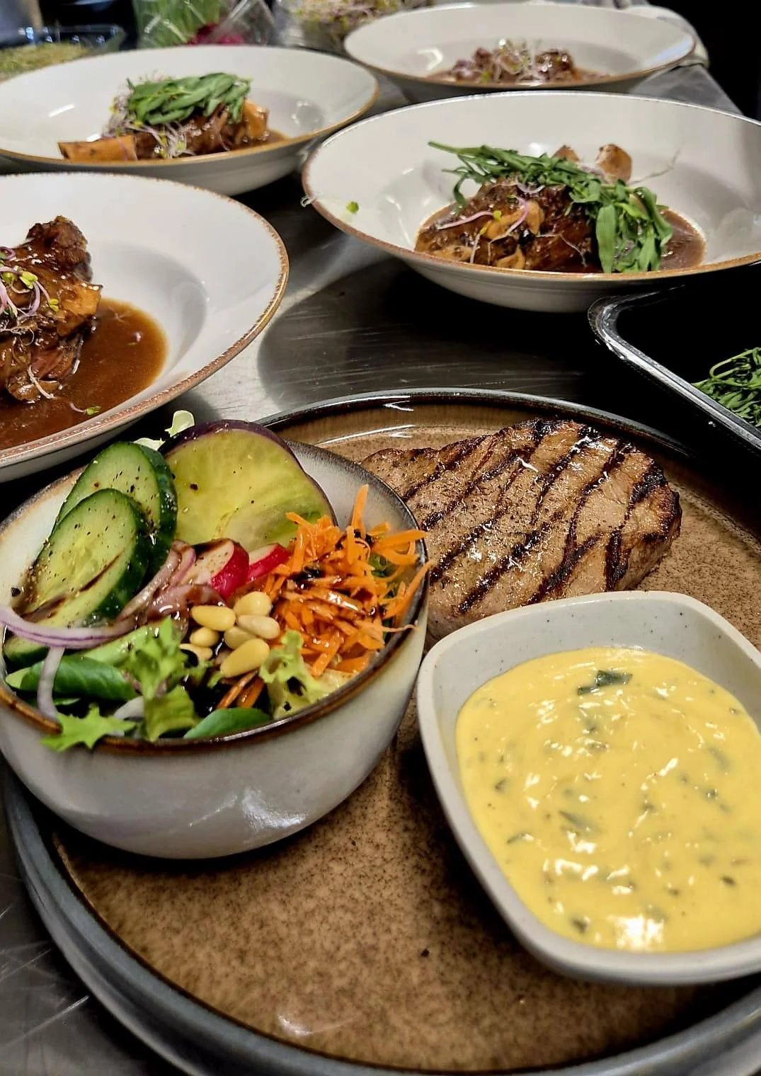 Assiette avec un steak grillé, salade avec avocat, carottes râpées et céleri, sauce moutarde, et plusieurs assiettes de viande avec légumes en arrière-plan  - Aftersport Clavier