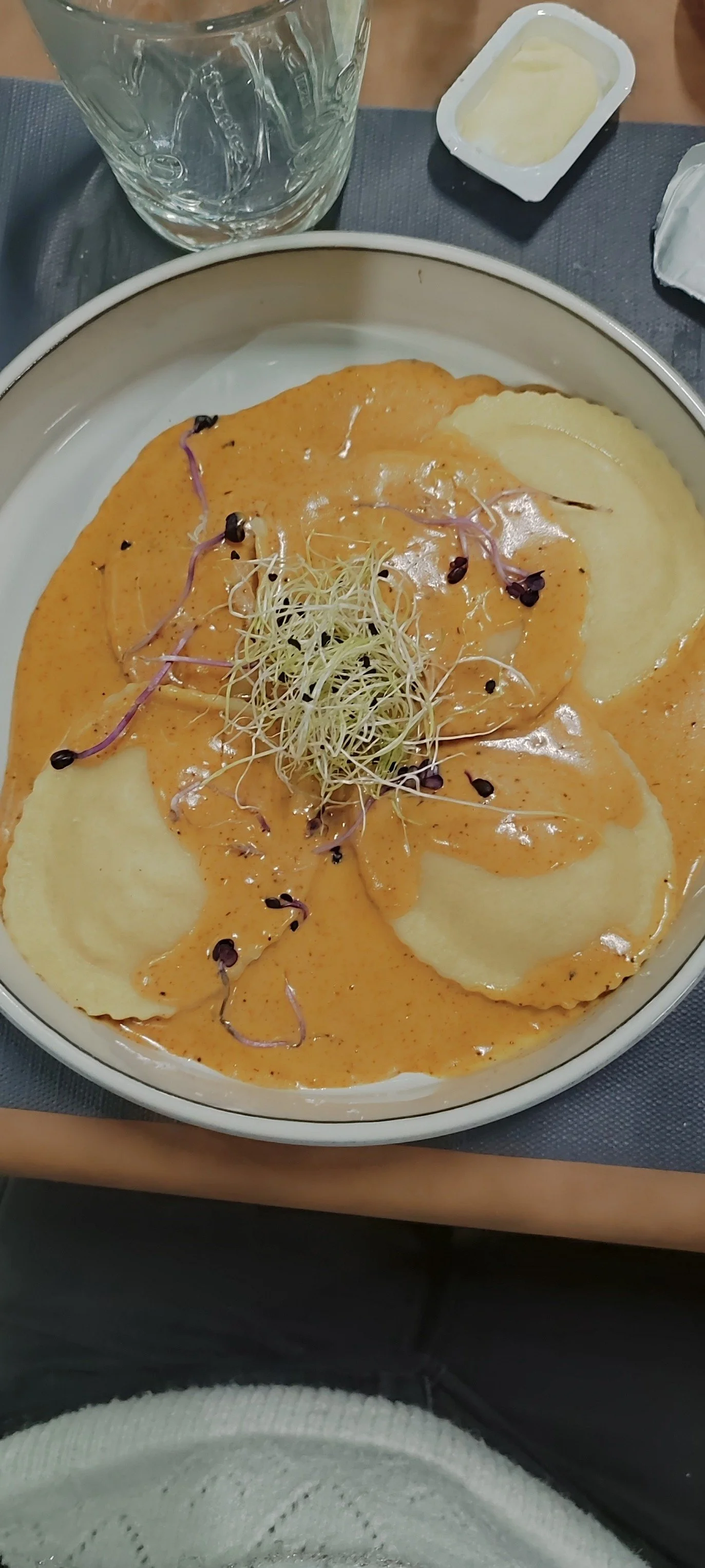 Assiette de raviolis avec une sauce à l'orange, garnie de pousses germées, accompagnée d'un verre d'eau et de beurre  - Aftersport Clavier