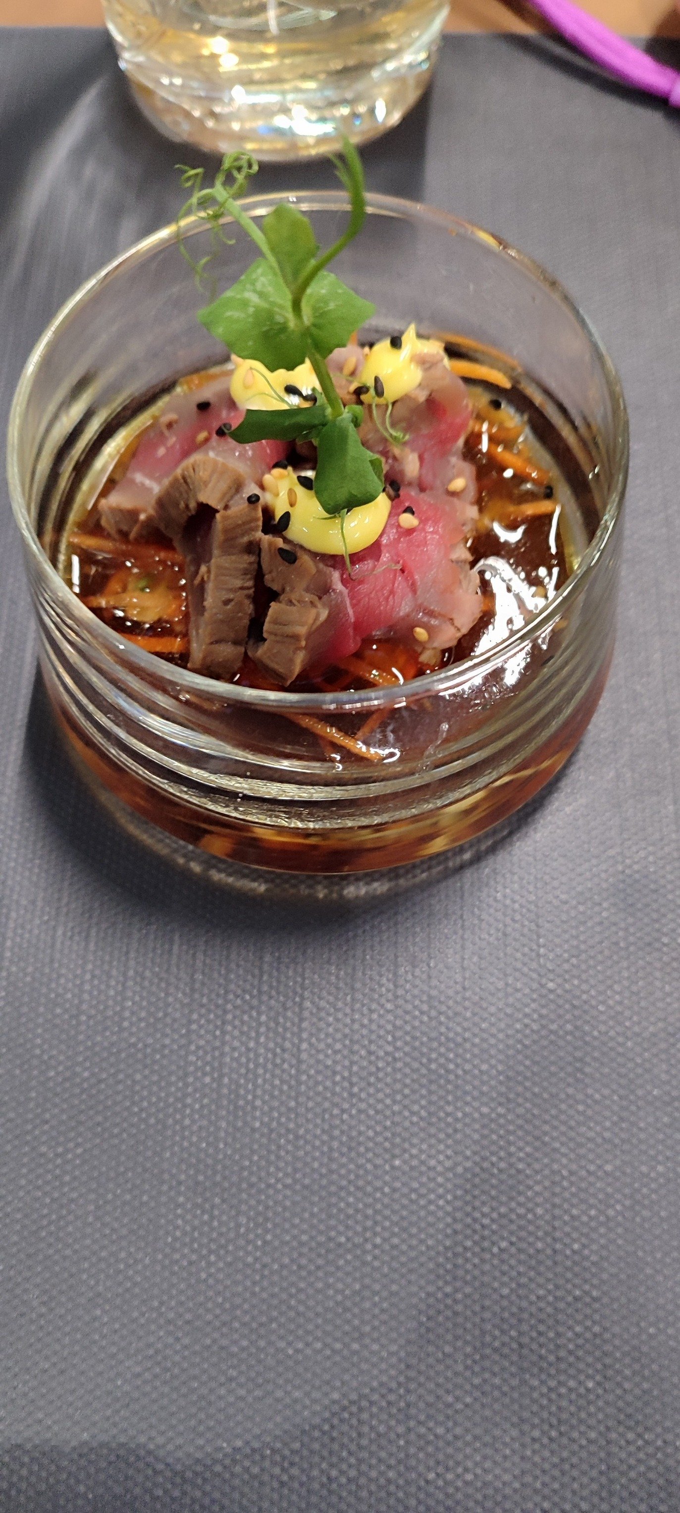 Plat présenté dans un petit bocal en verre, contenant de la viande de boeuf saignante, des petits morceaux de foie, des légumes julienne, de la sauce soja ou marinade, et décoré avec une petite pousse verte, une touche de mayonnaise, des graines de s