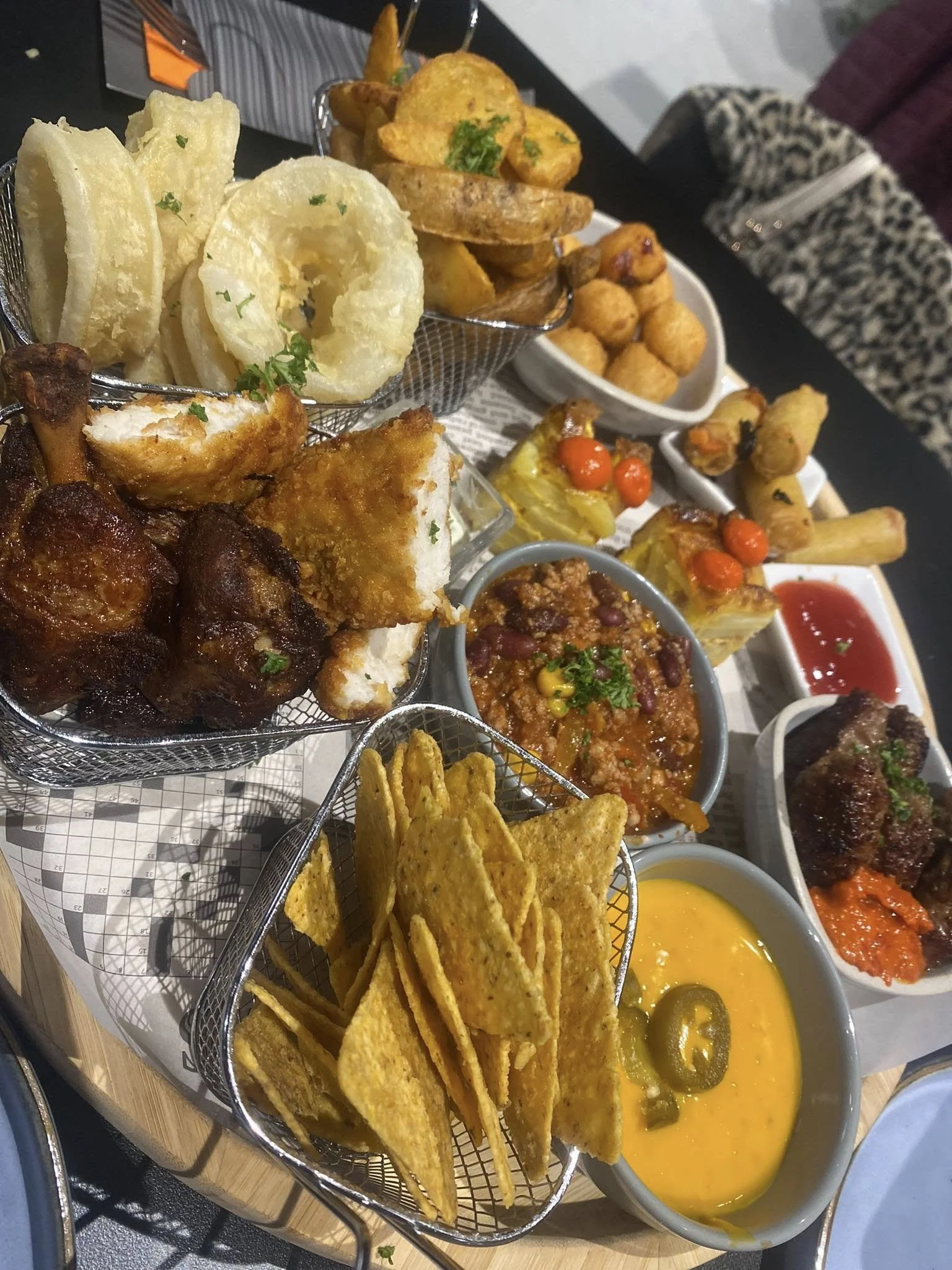 Plateau de divers snacks frits et sauces, incluant des rondelles d'oignon, des morceaux de poulet, des chips de tortilla, des pommes de terre rôties, des mini-tacos, des boulettes de viande, un bol de chili, et des sauces ketchup et barbecue  - After