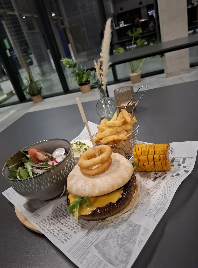 Un burger avec des oignons frits, des frites, un épi de maïs, de la salade et une poignée d'oignons frits, accompagné d'une salade et d'une boisson, sur une table dans un restaurant  - Aftersport Clavier