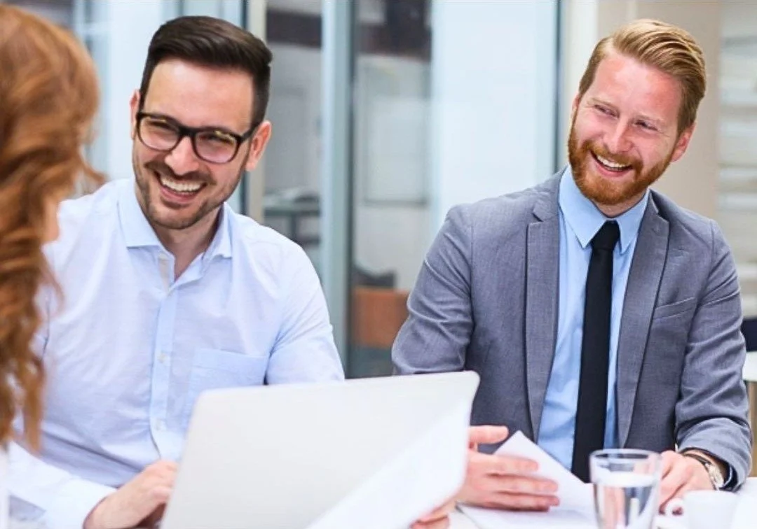 Four professionals are sitting at a table, engaging in conversation and smiling, with laptops and papers in front of them, in a modern office setting.