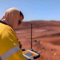 Person wearing a yellow jacket and beige headscarf using a surveying instrument outdoors on a red desert landscape.