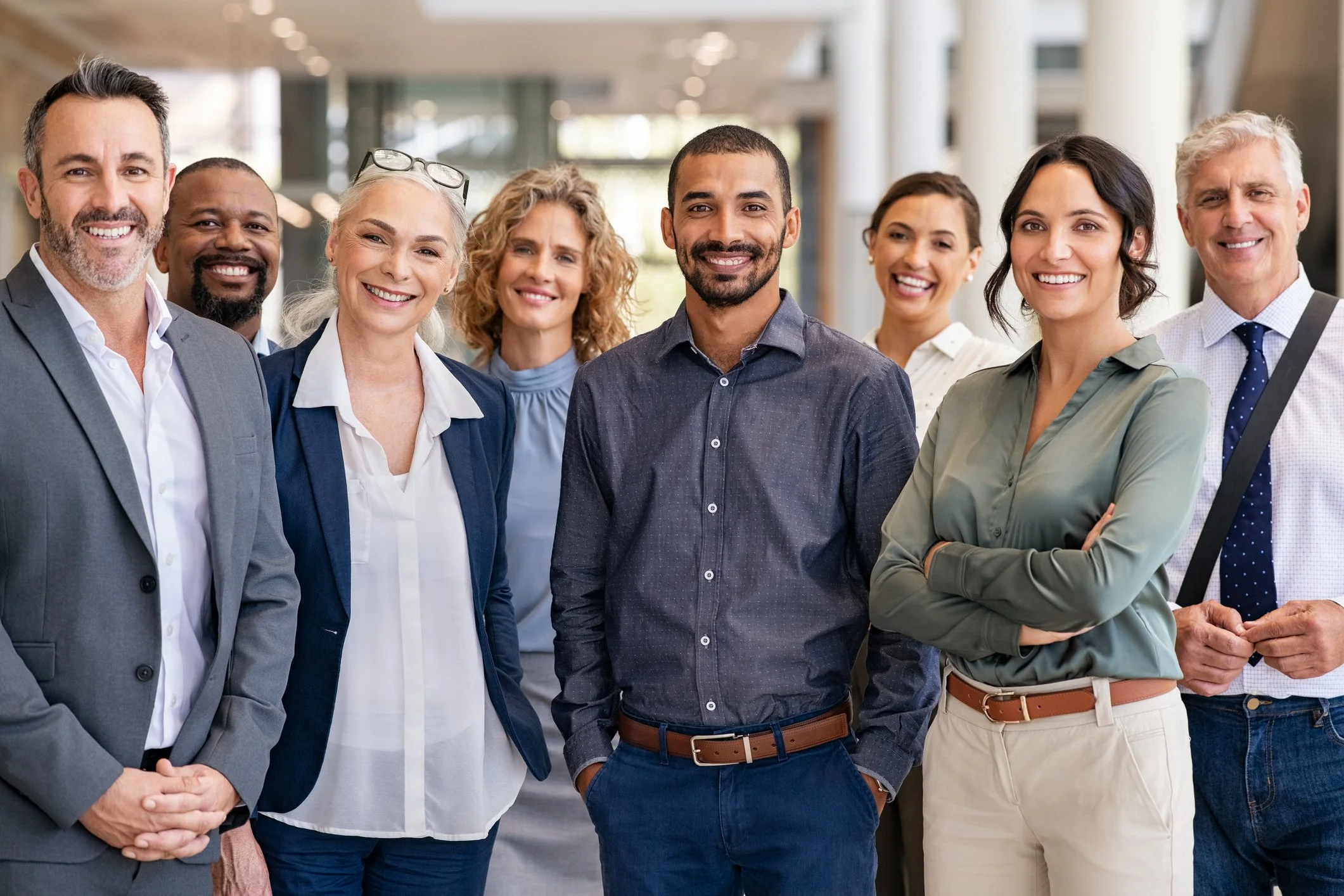 A diverse group of eight professional people standing together indoors in a modern office building, smiling at the camera.