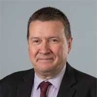 Portrait of a middle-aged man with short brown hair, wearing a dark suit, light shirt, and patterned tie, against a neutral background.