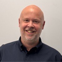 Headshot of a smiling bald man with a beard, wearing a dark collared shirt, against a plain light-colored background.