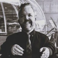 A smiling man with a beard and mustache, wearing a dark suit and tie, sitting outdoors with a background of a glass structure and trees.