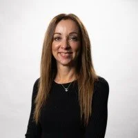A woman with long brown hair wearing a black top, smiling against a plain white background.