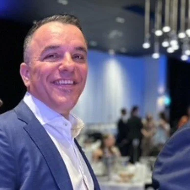 Smiling man in a suit at a formal event with a crowd and tables in the background.