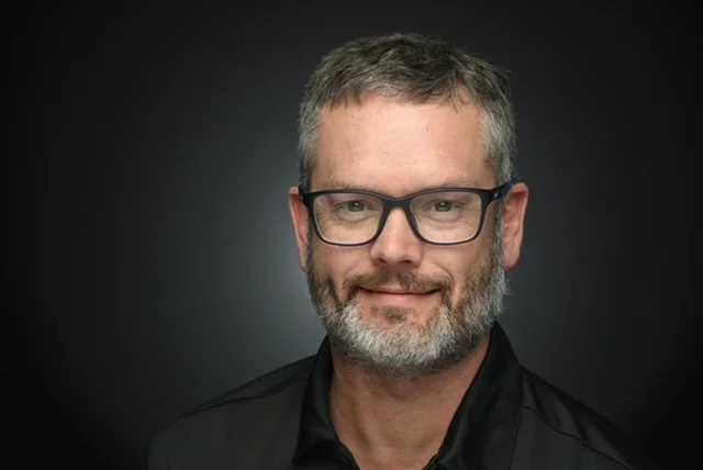 Headshot of a middle-aged man with short gray hair, glasses, a beard, and a black jacket against a dark background.