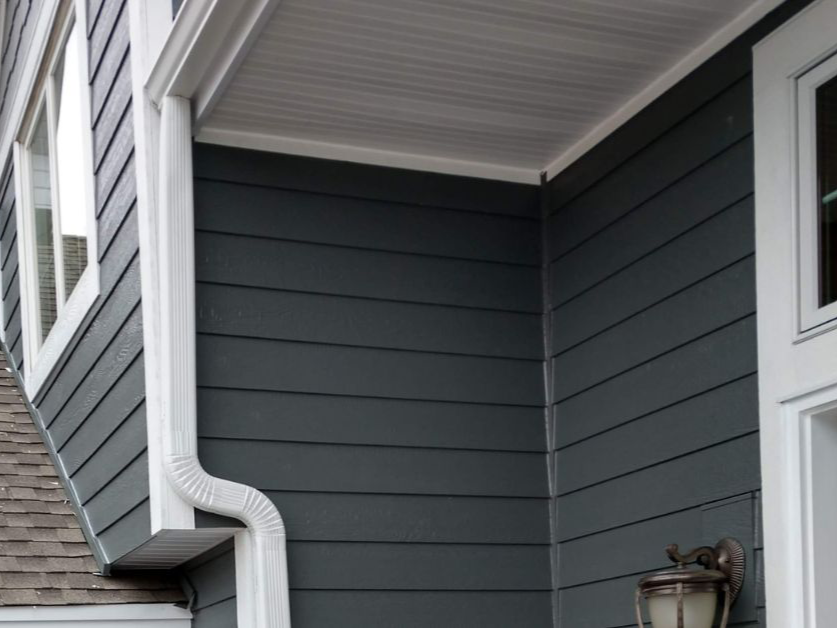 Close-up of the corner of a house showing blue horizontal siding, a white gutter, and a white soffit under the roof overhang.