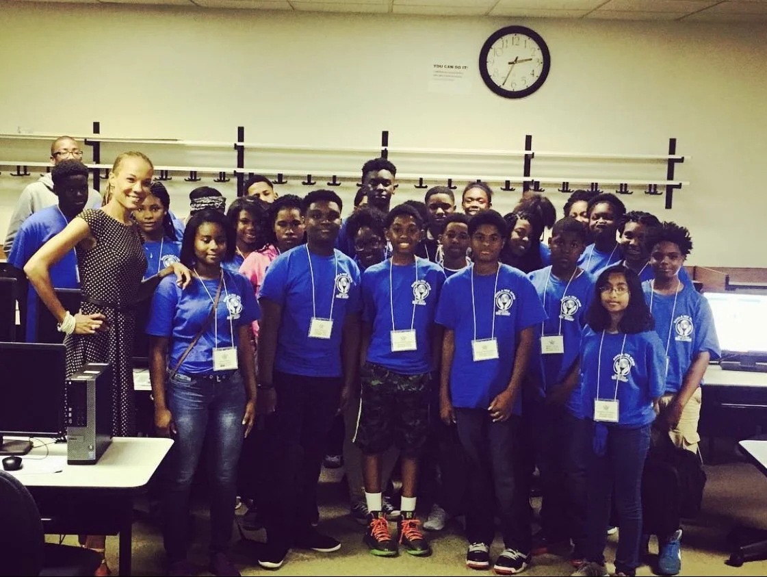 Group of young students and a woman teacher in a classroom, some wearing blue shirt with an emblem, posing for a photo with a clock and computers in the background.