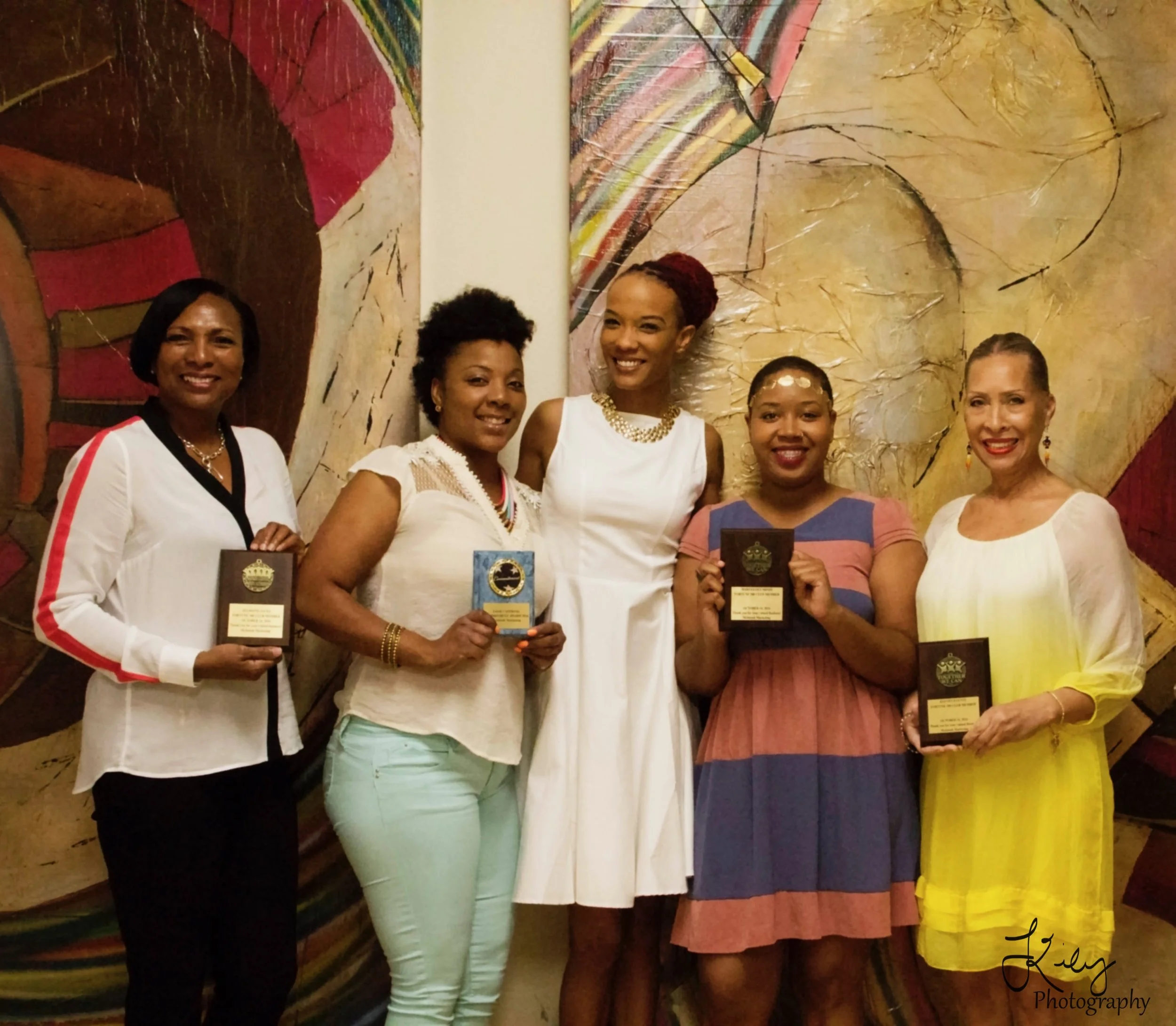 Five women standing side by side, holding plaques or awards, smiling in front of an abstract art background.