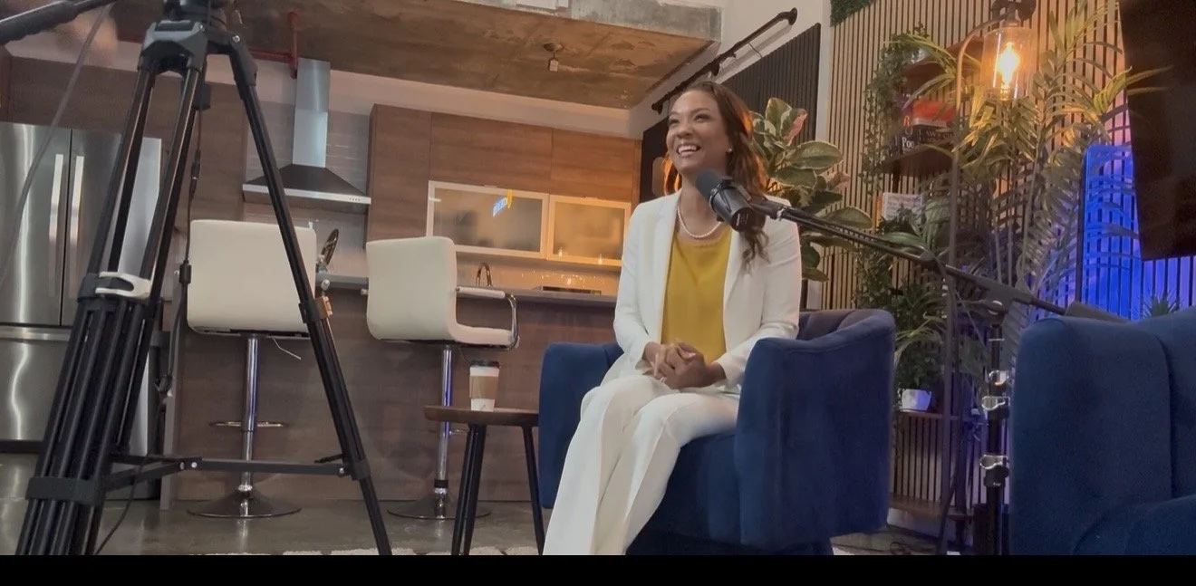 A woman sitting on a blue armchair, smiling and speaking into a microphone, in a modern, cozy indoor setting with plants, lights, and a kitchen in the background.