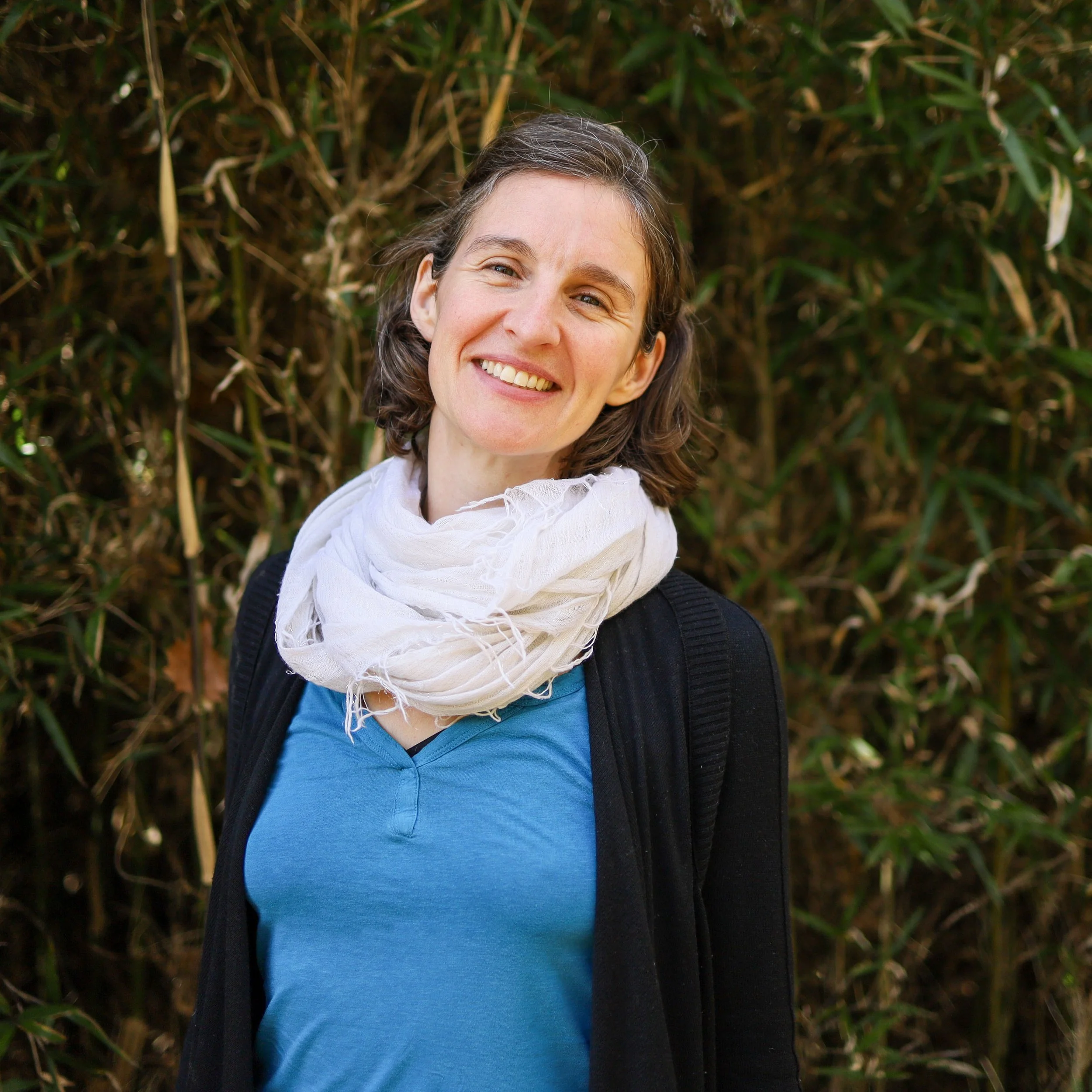 A woman with shoulder-length brown hair, smiling, wearing a blue shirt, white scarf, and black jacket, standing in front of green foliage.