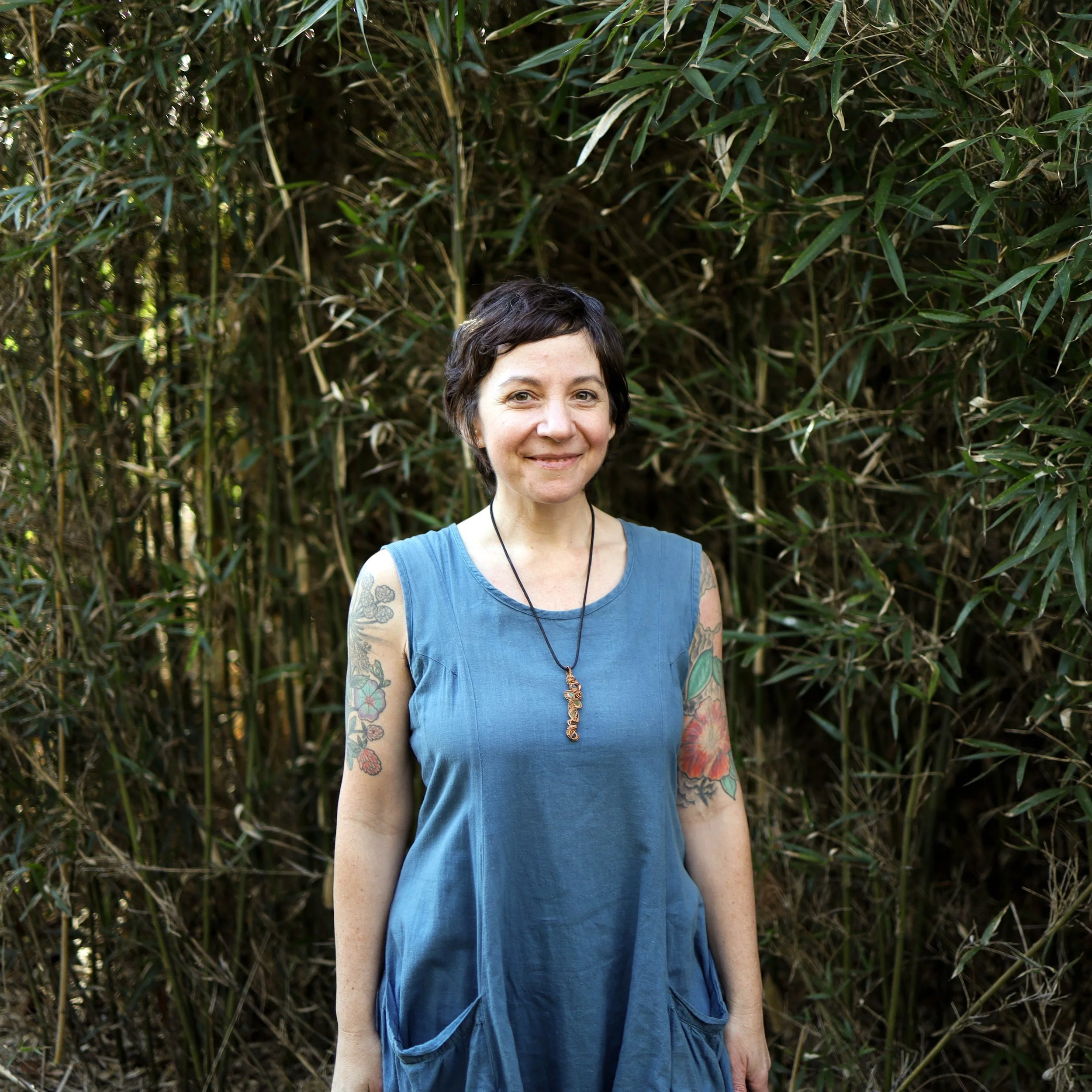 Woman with short dark hair, wearing a blue sleeveless dress and a necklace, standing outdoors in front of dense green foliage, smiling.