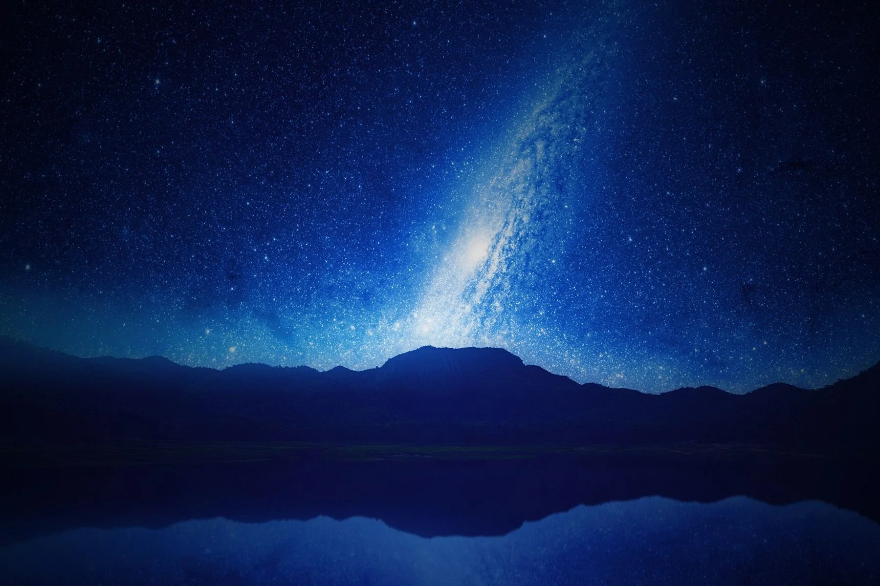 Nighttime landscape with a Milky Way galaxy stretching across the sky, mountain silhouette in the foreground, and a calm body of water reflecting the starry sky.