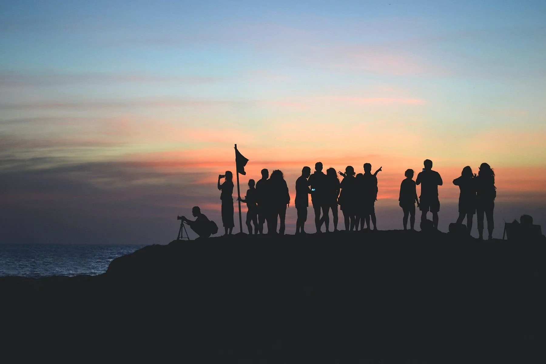 Group of people silhouetted at sunset on a rocky coastline, some taking photos, holding a flag, or looking at the ocean.