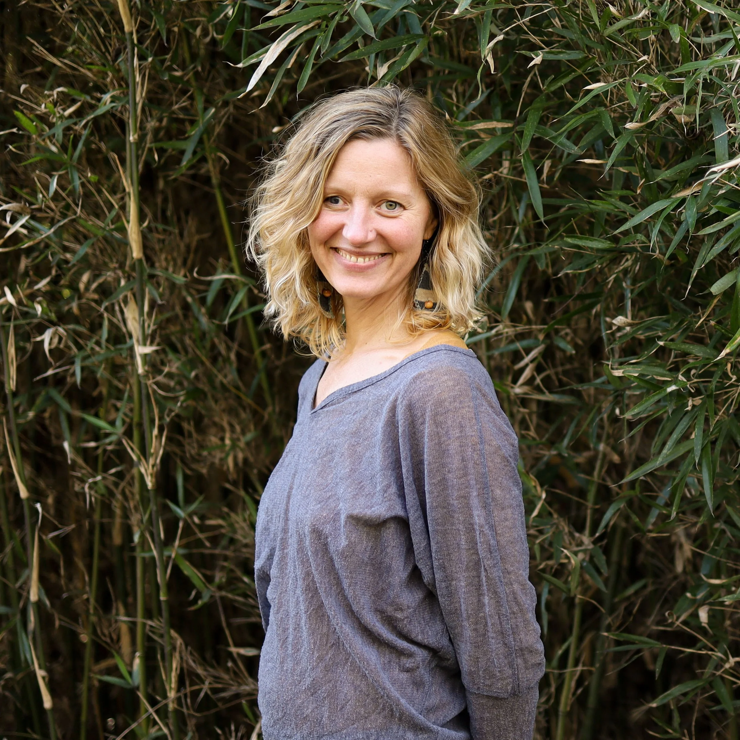 A woman with blonde, wavy hair smiling at the camera, standing outdoors in front of green foliage.