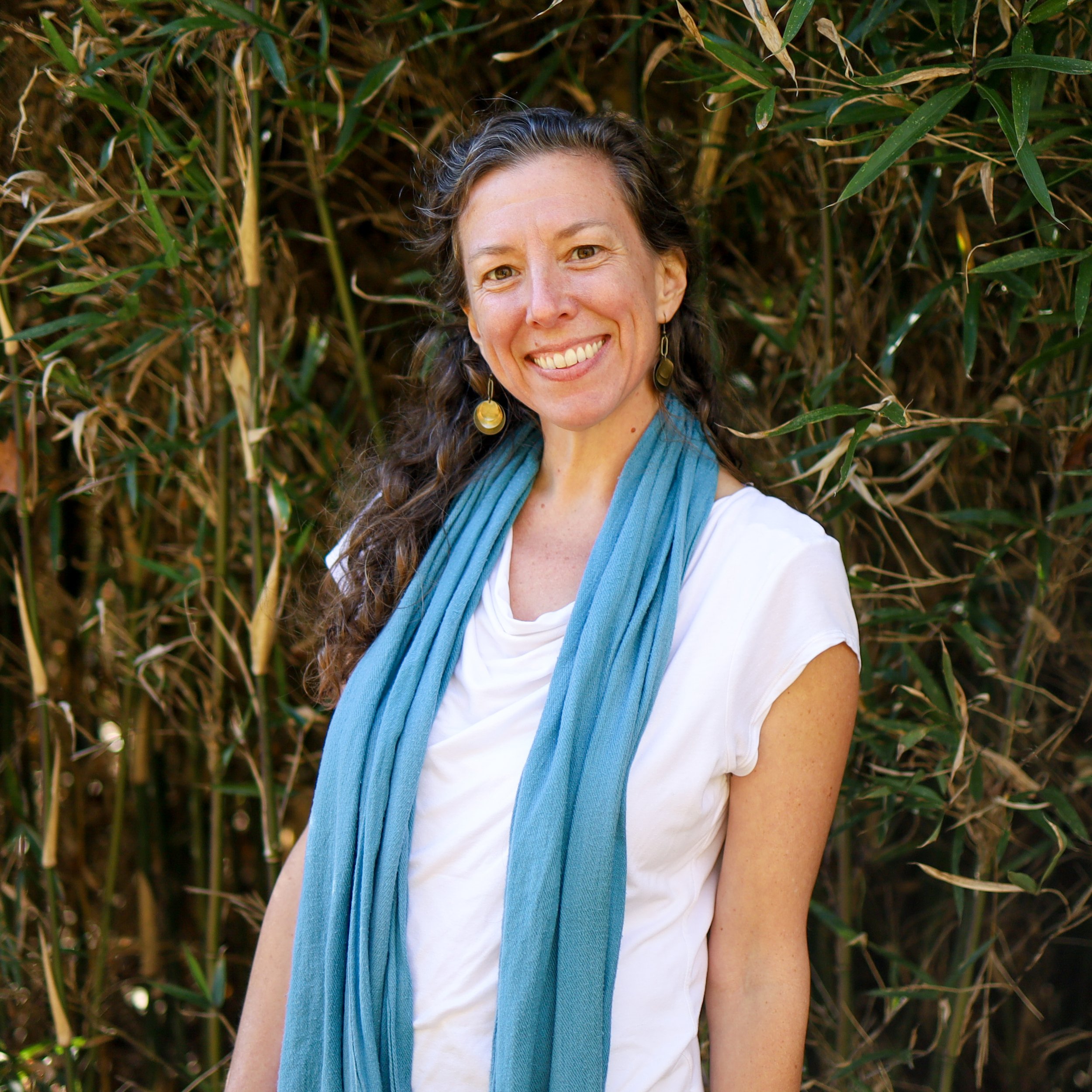 A woman with long, wavy brown hair, smiling, wearing a white top, gold earrings, and a light blue scarf, standing outdoors in front of green bamboo stalks.